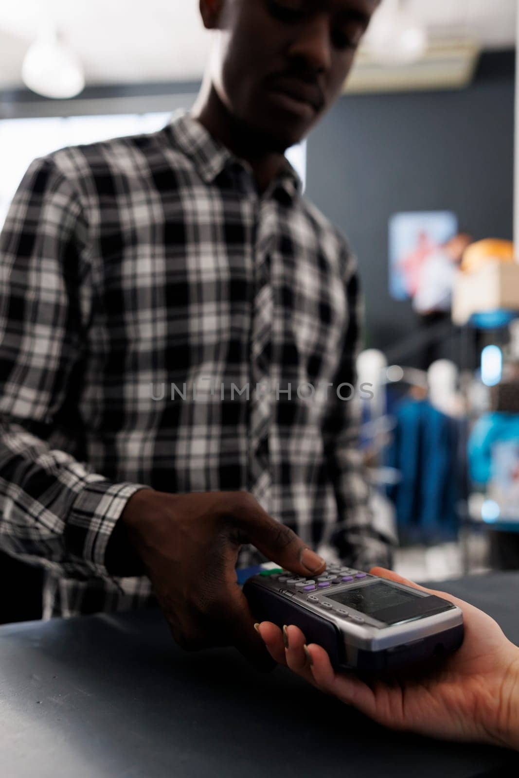 African american customer typing credit card details on pos to pay for stylish clothes, making contactless payment at store counter. Shopaholic man buying fashionable items for casual wear in boutique