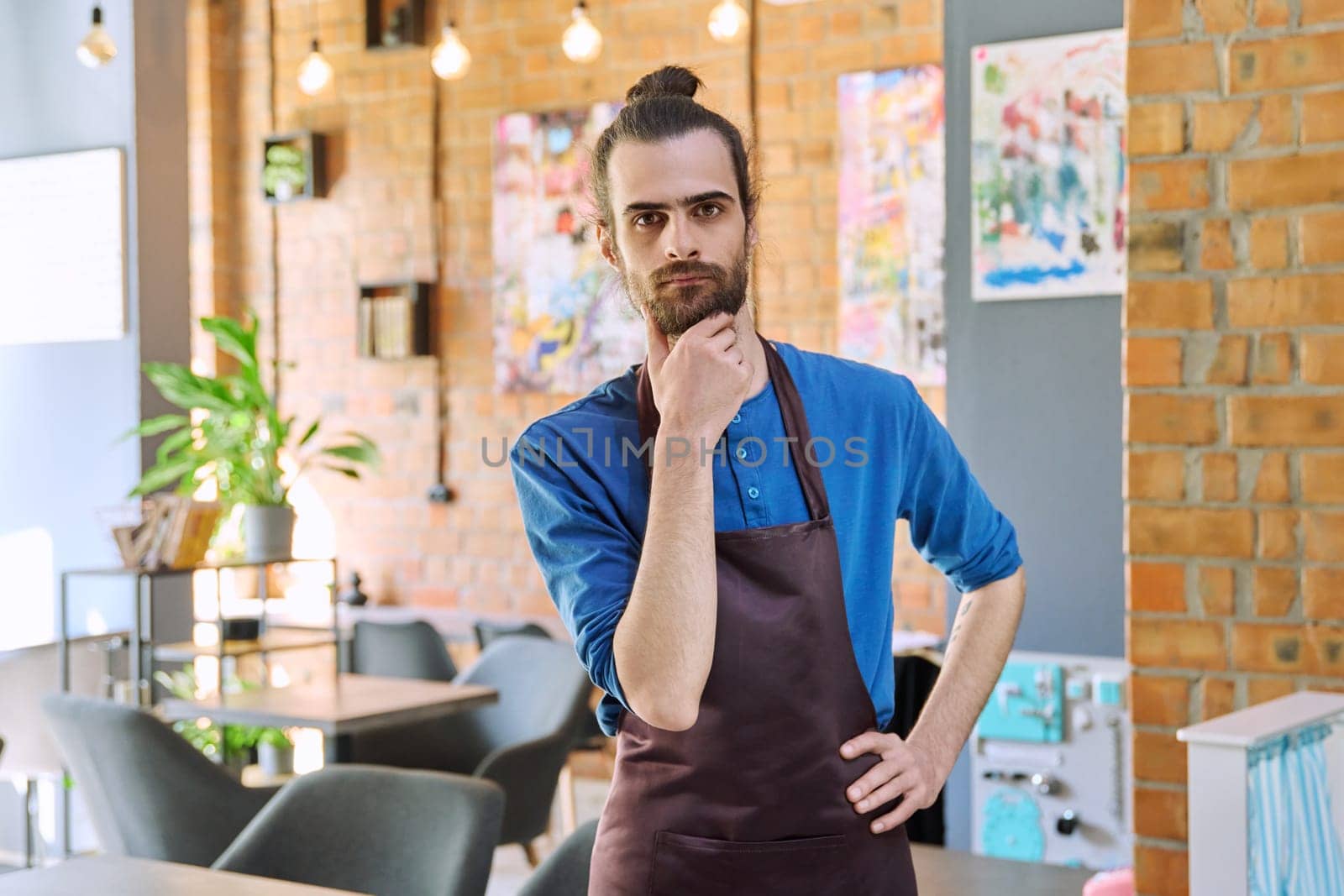 Confident successful young man service worker owner in apron posing looking at camera in restaurant cafeteria coffee pastry shop interior. Small business staff occupation entrepreneur work