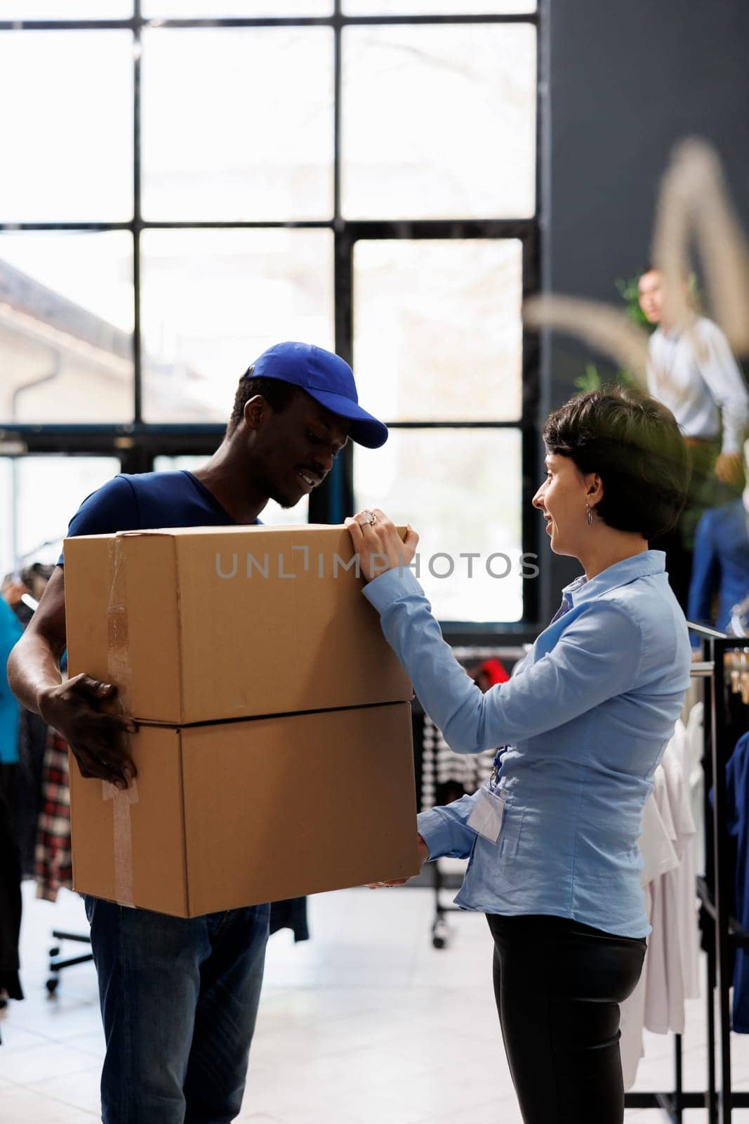 Store manager discussing client orders detalis with african american courier in modern boutique. Man wearing delivery uniform, holding cardboard boxes with stylish clothes in shopping centre