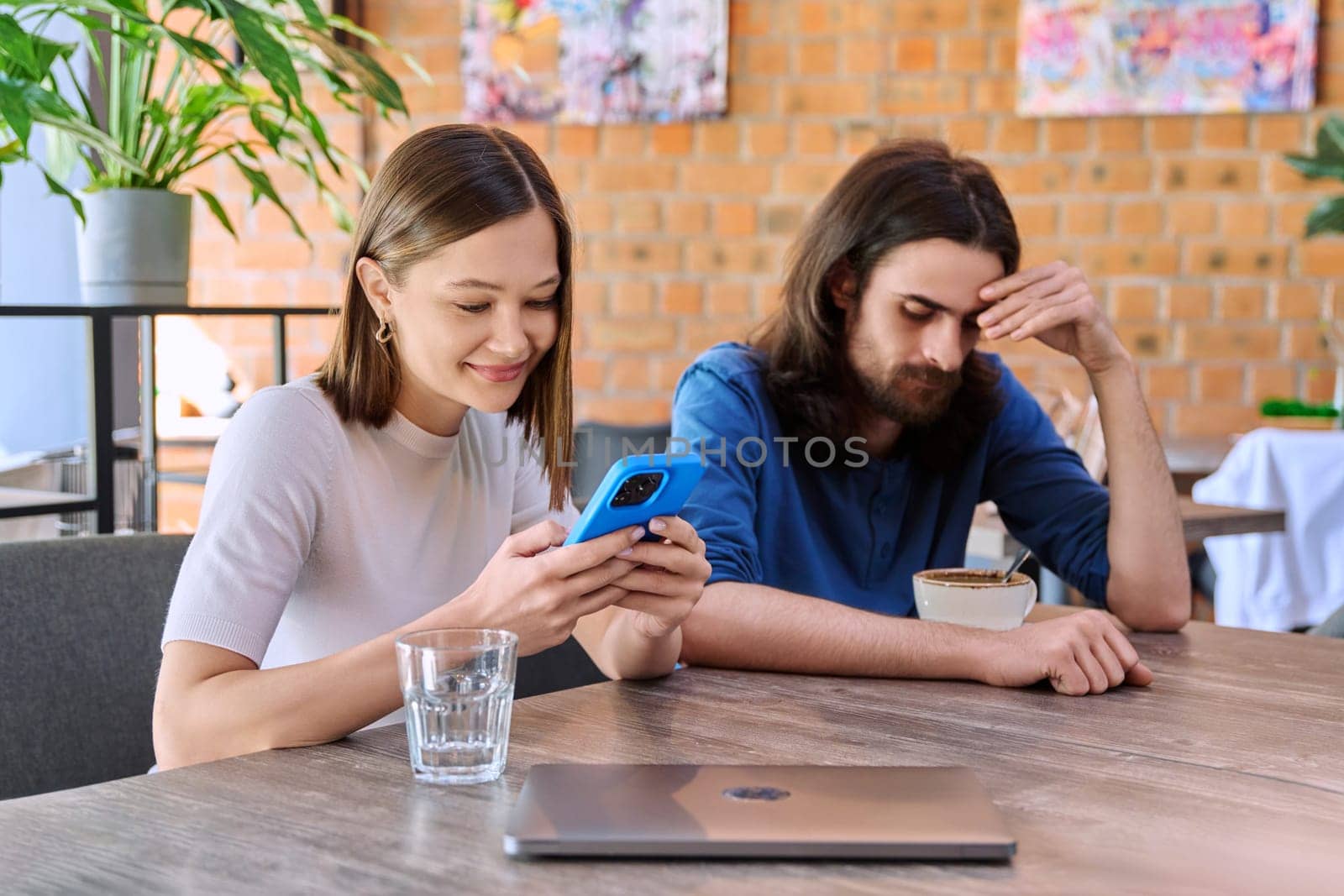 Young handsome people friends, couple man and woman relaxing together in a cafe, using smartphone, drinking coffee. Lifestyle, leisure, youth concept