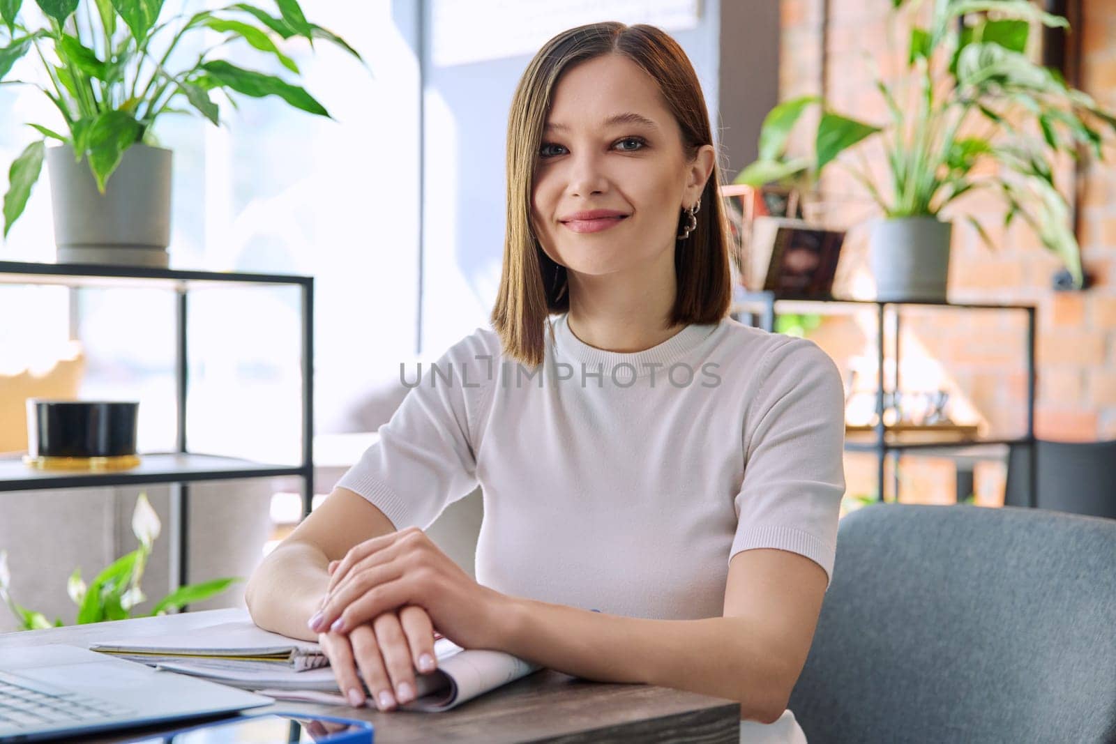 Portrait of beautiful smiling young woman sitting at table in coworking cafe, stylish pretty female 20s student looking at camera. Youth, beauty, university college students, lifestyle concept