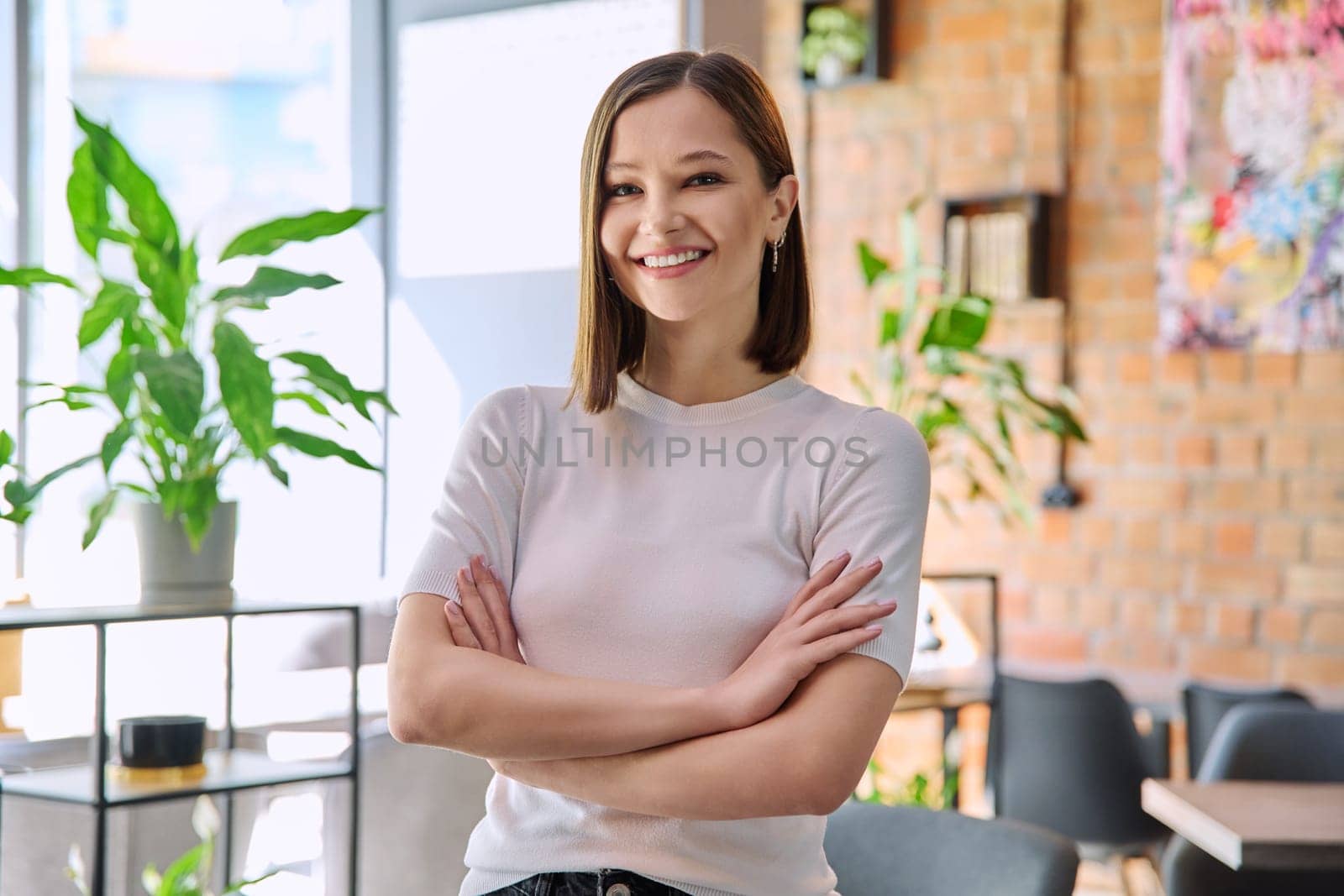 Portrait of young confident beautiful woman with crossed arms looking at camera in coworking cafe. 20s female university college student, businesswoman, freelancer employee, young specialist