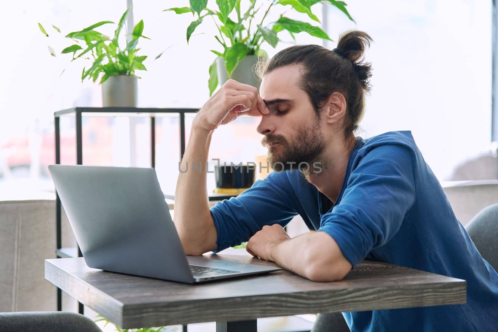 Young tired man working with laptop computer having headache migraine fatigue, holding hands on face. Depression, stress, problems, difficulties with business work, mental health