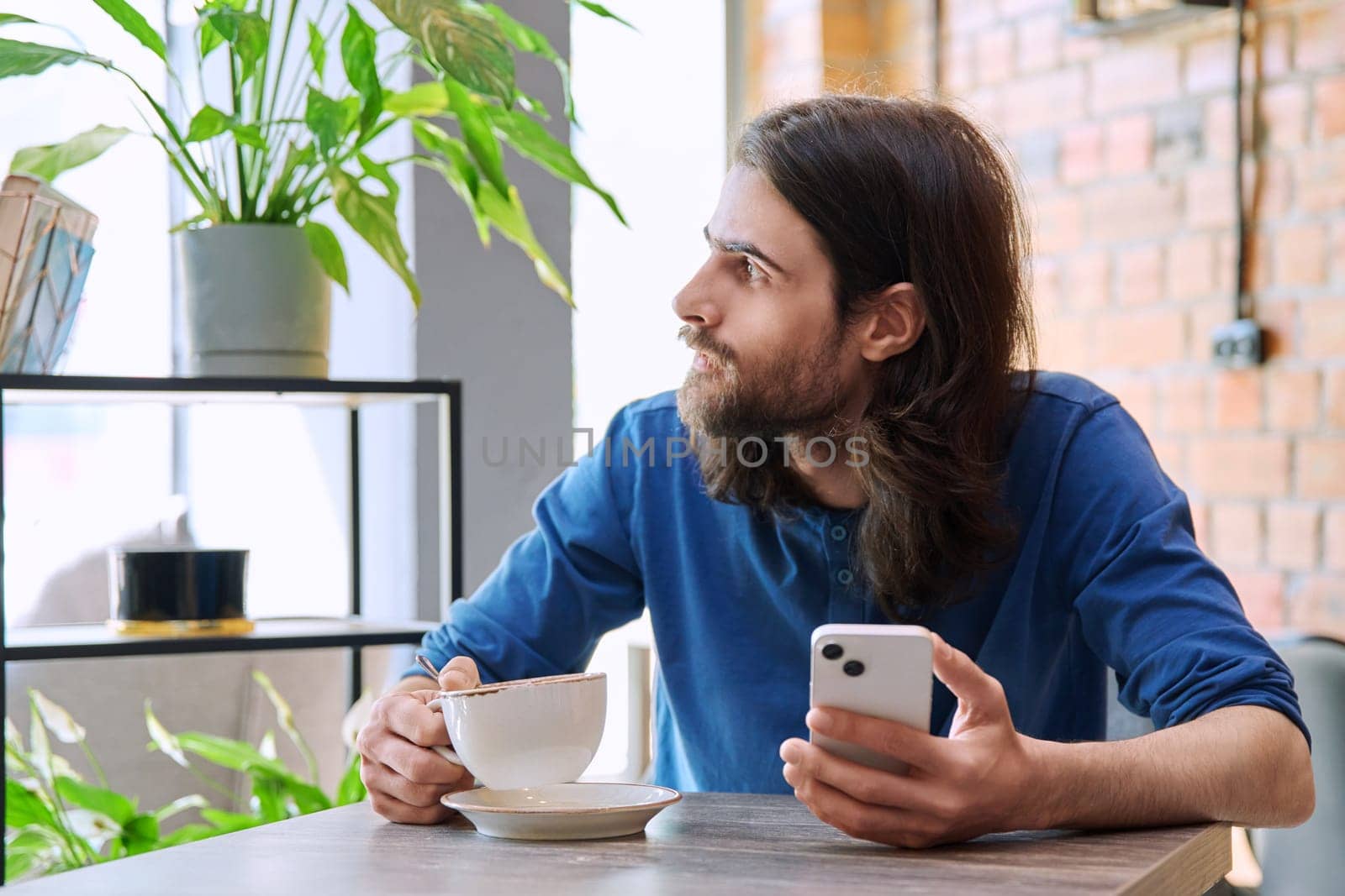 Young handsome man using smartphone, drinking cup of coffee, sitting in cafe by VH-studio