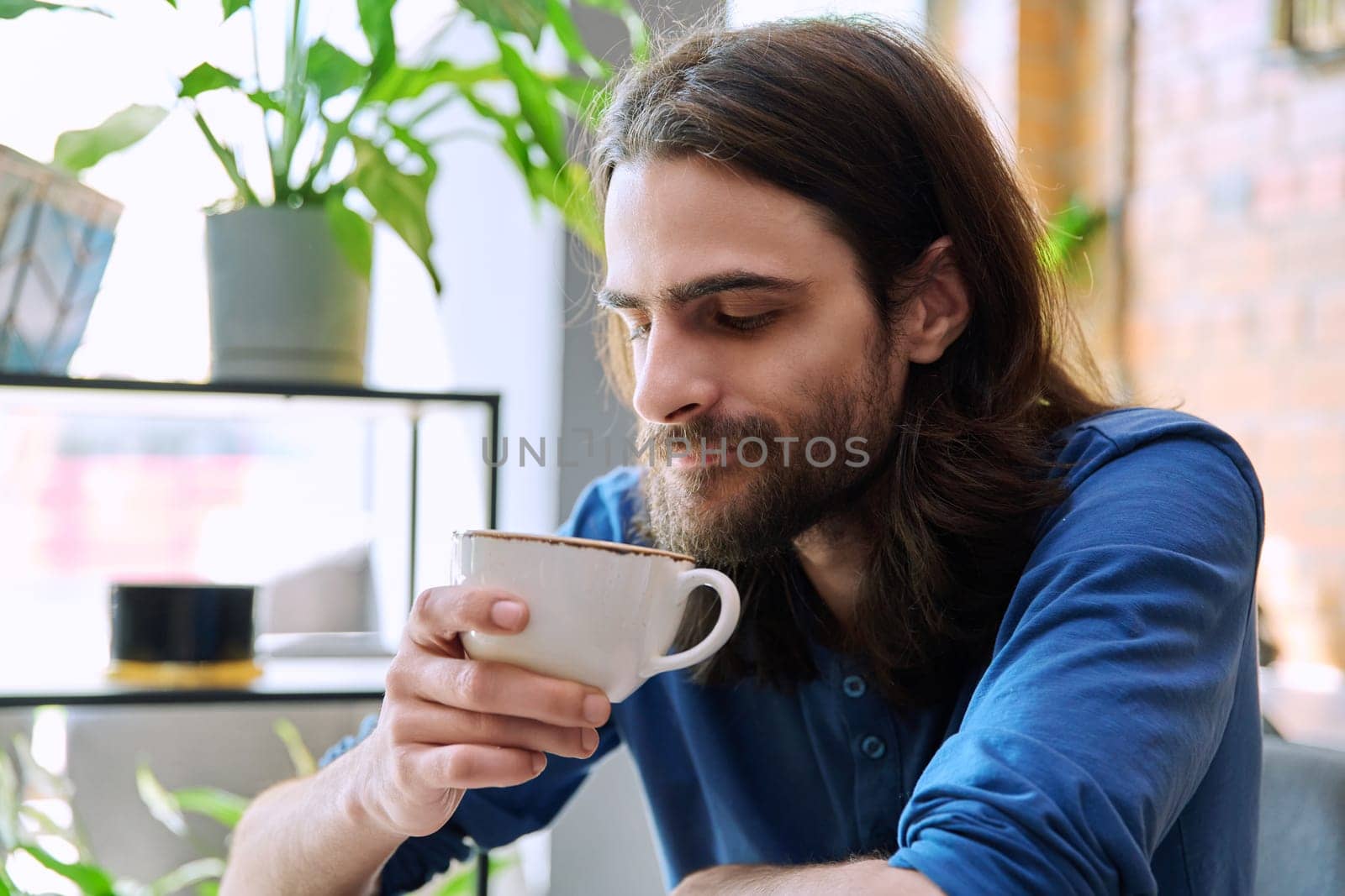 Close up of young handsome man holding drinking cup of coffee while sitting in coffee shop restaurant. Lifestyle, leisure, relaxation, coffee drinks, people concept