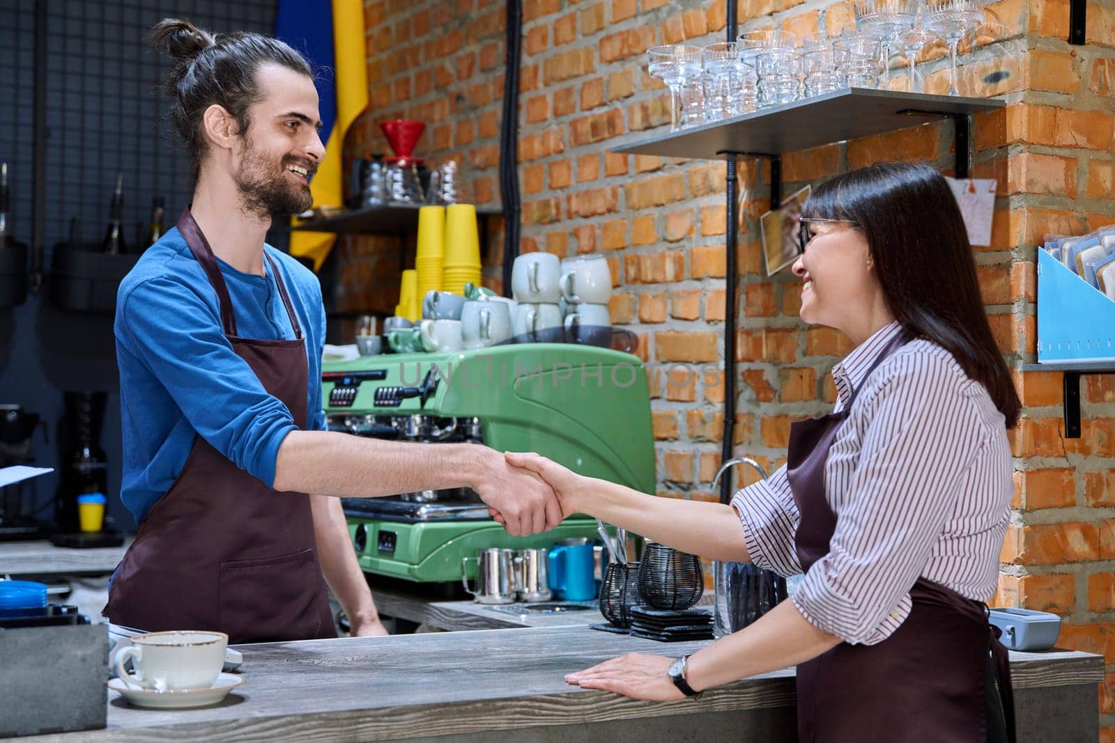 Young man in apron shaking hands with woman owner of coffee shop by VH-studio