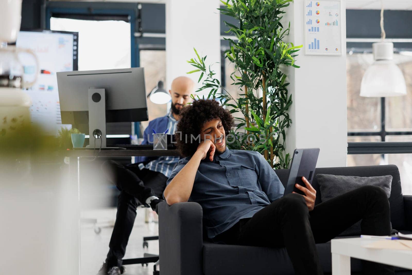 Start up business company manager watching video on digital tablet during break in office. Arab young entrepreneur browsing social media and relaxing on couch in coworking space