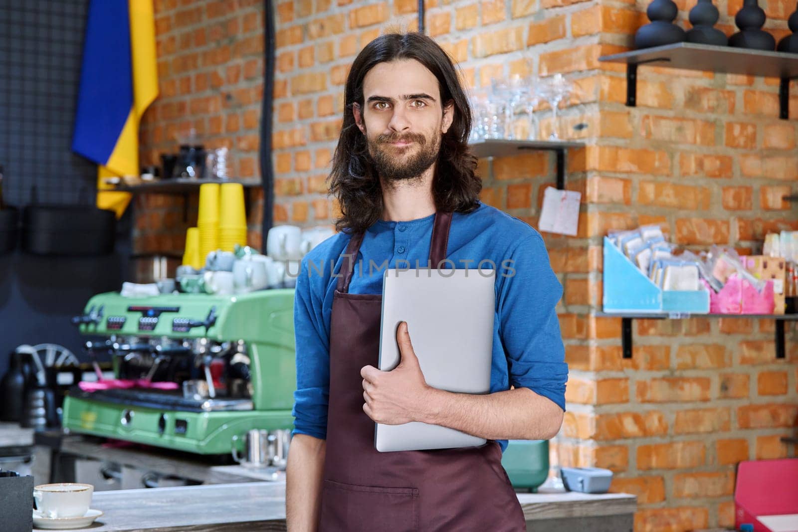 Young man in apron, food service worker, small business owner entrepreneur holding laptop looking at camera near counter of coffee shop cafe cafeteria. Staff, occupation, successful business, work