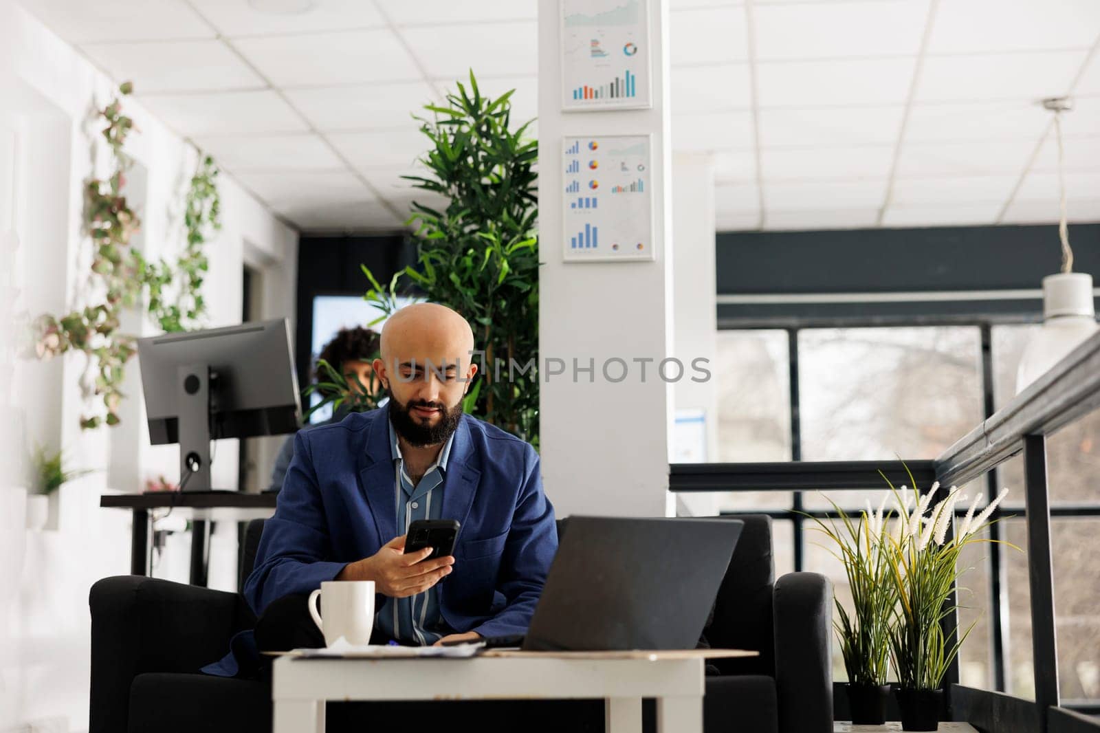Business arab professional texting on smartphone while working on laptop in start up office. Entrepreneur searching financial information on mobile phone in green open space