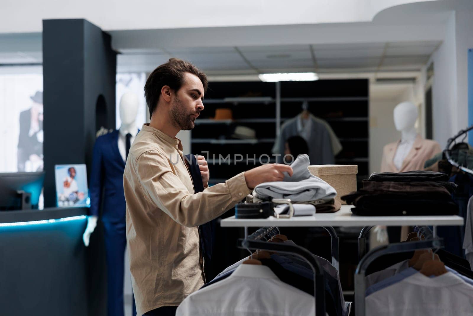 Young man shopping for trendy menswear, browsing through shelf in clothing store. Fashion boutique customer choosing apparel and accessory, searching for size and examining style