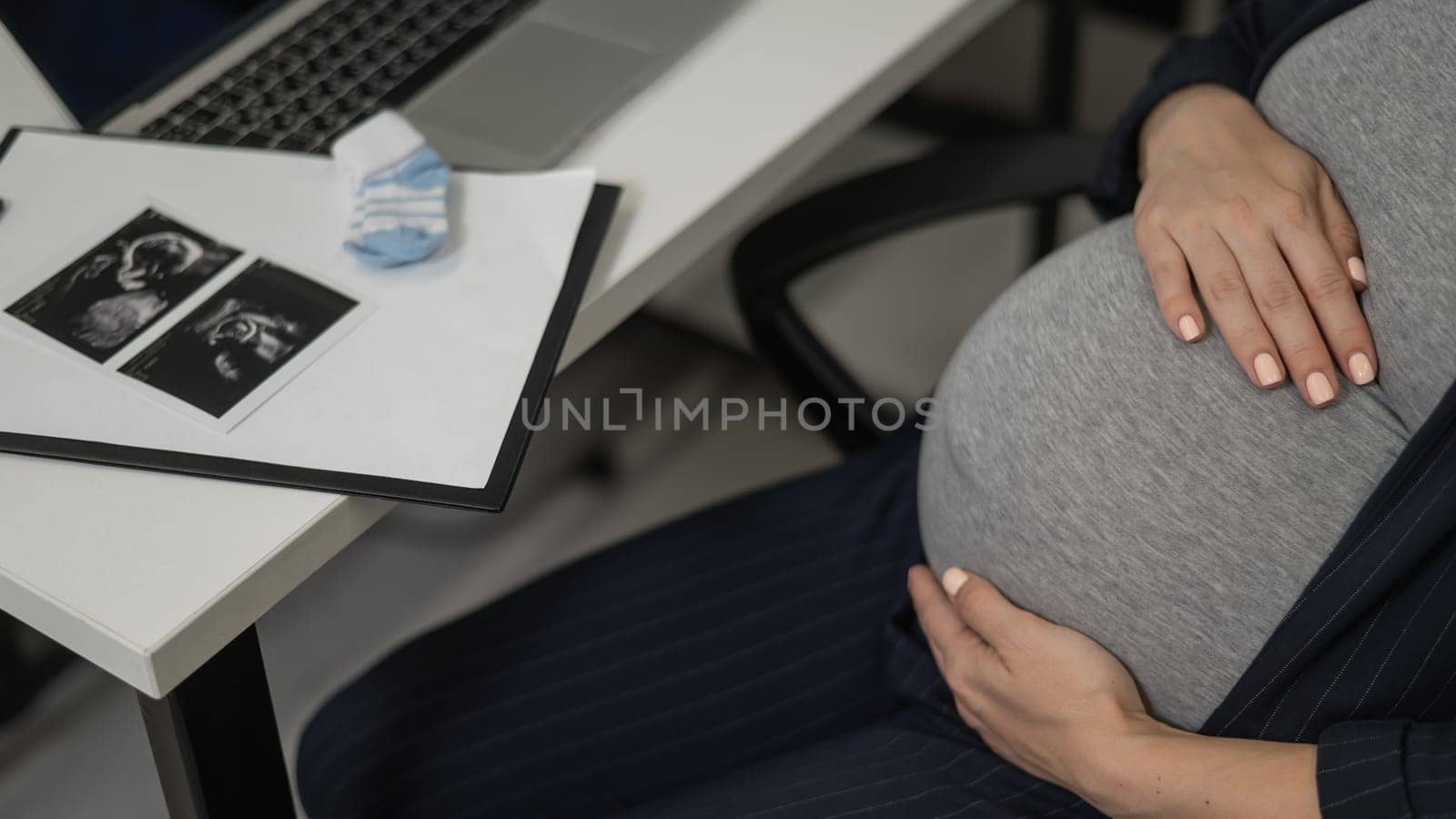 A pregnant woman works on a laptop in the office and looks at a photo from an ultrasound scan of the fetus. Belly close-up. by mrwed54