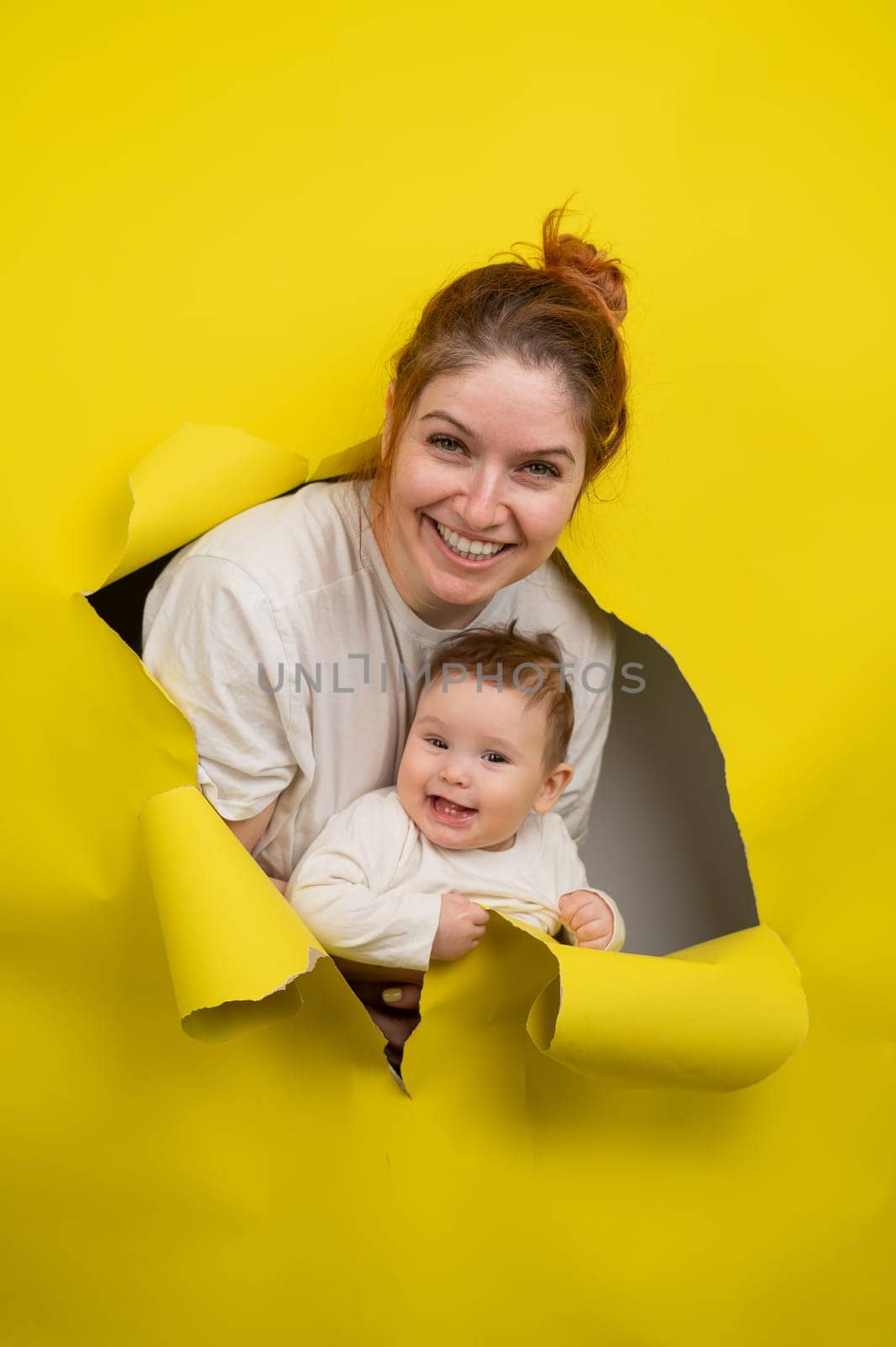 Cheerful Caucasian woman with little son rips and leans out through yellow cardboard background. Vertical photo