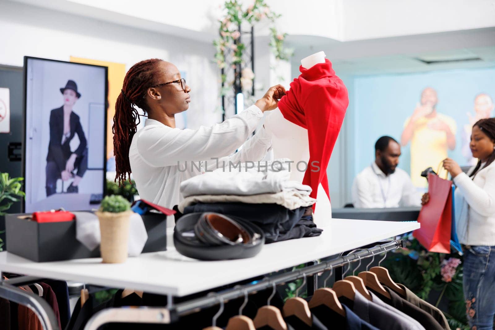 African american woman dressing mannequin in red pullover in clothing store. Shopping mall assistant putting female casual blouse on dummy model to display apparel for sale