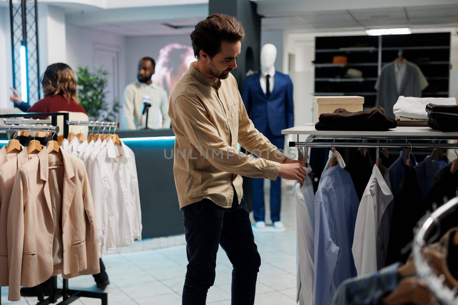 Young man customer examining apparel options in clothing store department while choosing formal wear. Buyer checking shirts while exploring rack in shopping mall showroom