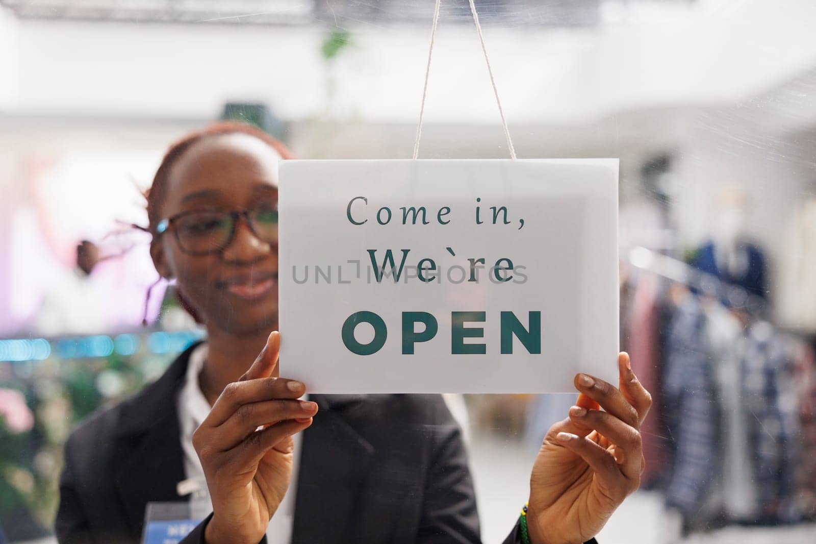 Fashion boutique shop smiling employee hanging we are open sign on store front door. Young african american clothing mall department manager holding signboard with come in message