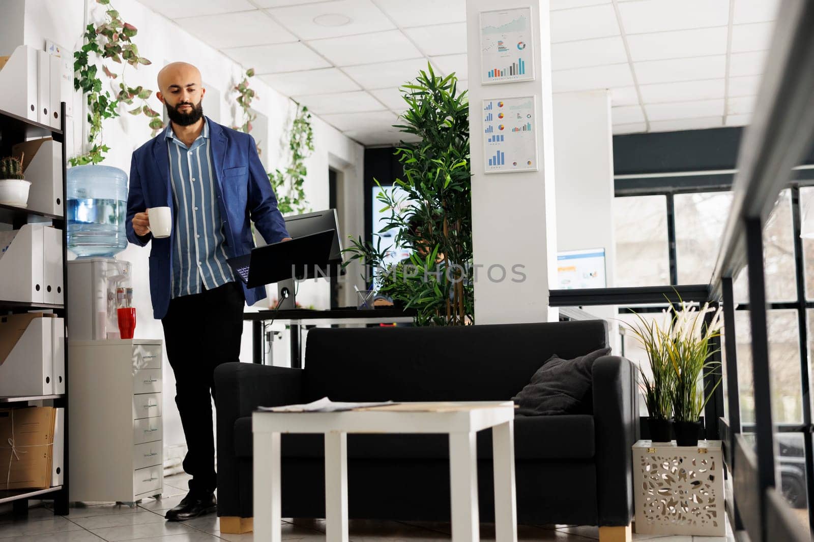 Arab man manager drinking coffee in start up business modern office. Young entrepreneur holding tea mug and laptop while working at strategy plan in coworking green open space