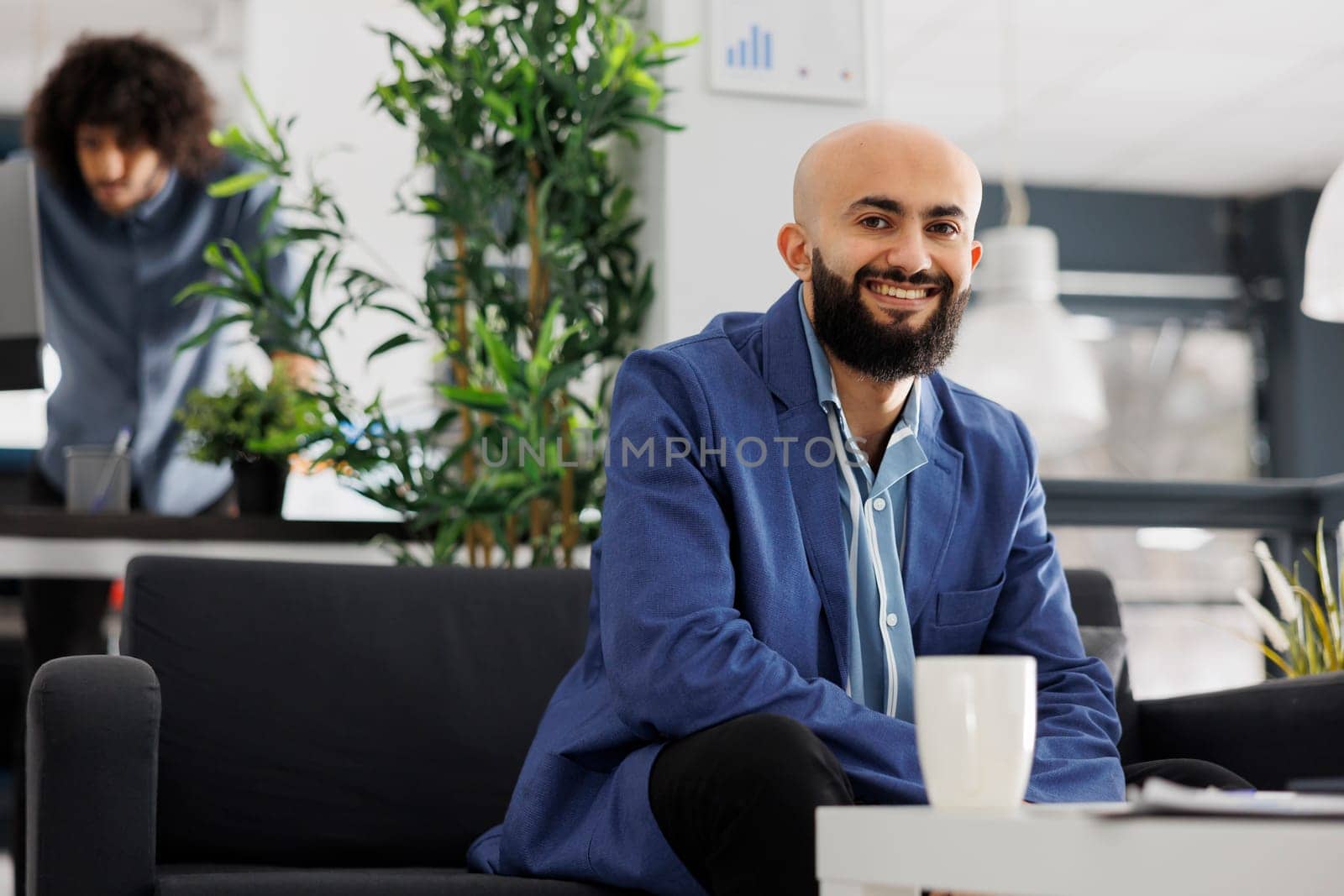 Smiling arab business entrepreneur looking at camera while sitting on couch in office. Start up company successful executive manager wearing suit portrait in coworking space