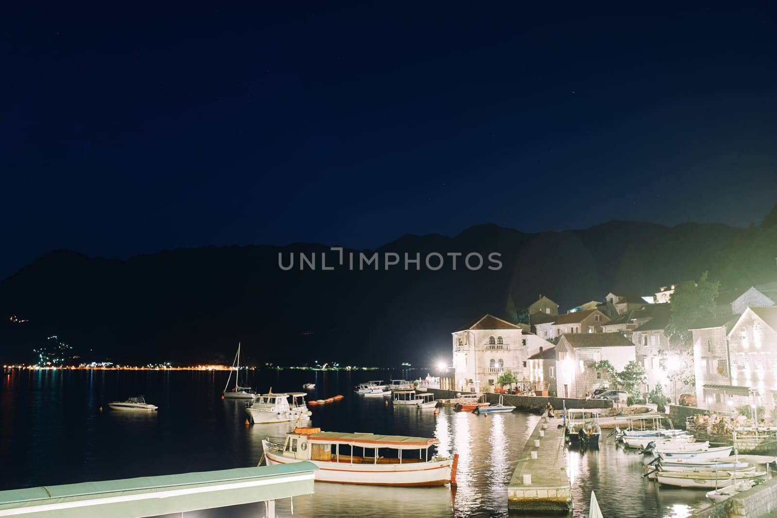 Fishing boats are moored at an illuminated pier at night near the coast of an ancient town. Perast. Montenegro by Nadtochiy