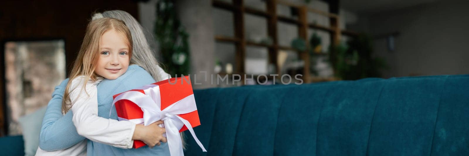 Happy little granddaughter and european old woman hugging, holding box with gift, celebrate birthday in living room. Love and family relationship, birthday, emotions and lifestyle at home, copy space