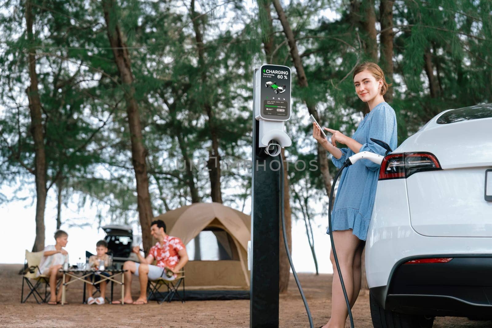 Outdoor adventure and family vacation camping at sea travel by eco friendly car. Woman or mother check car's battery with smartphone while charging EV car frin charging station in campsite. Perpetual