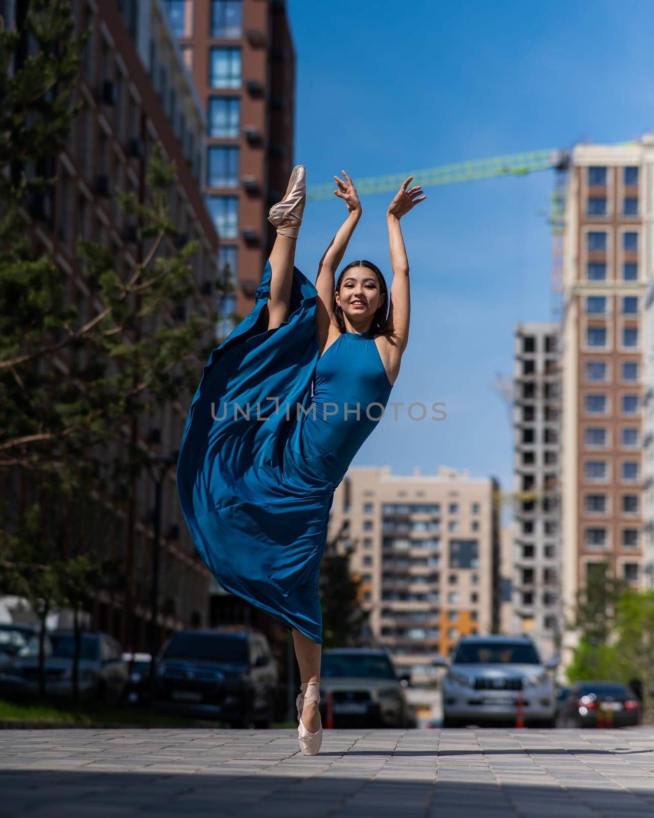 Beautiful Asian ballerina in blue dress posing in splits outdoors. Urban landscape. Vertical photo. by mrwed54