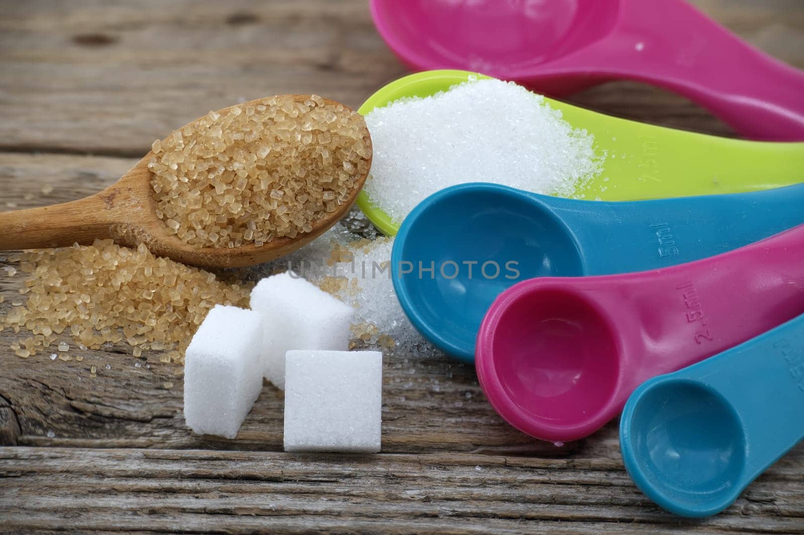 Brown cane sugar and white sugar in colorful plastic measuring spoons spilled around on a rustic wooden surface, measuring utensils in a kitchen setting