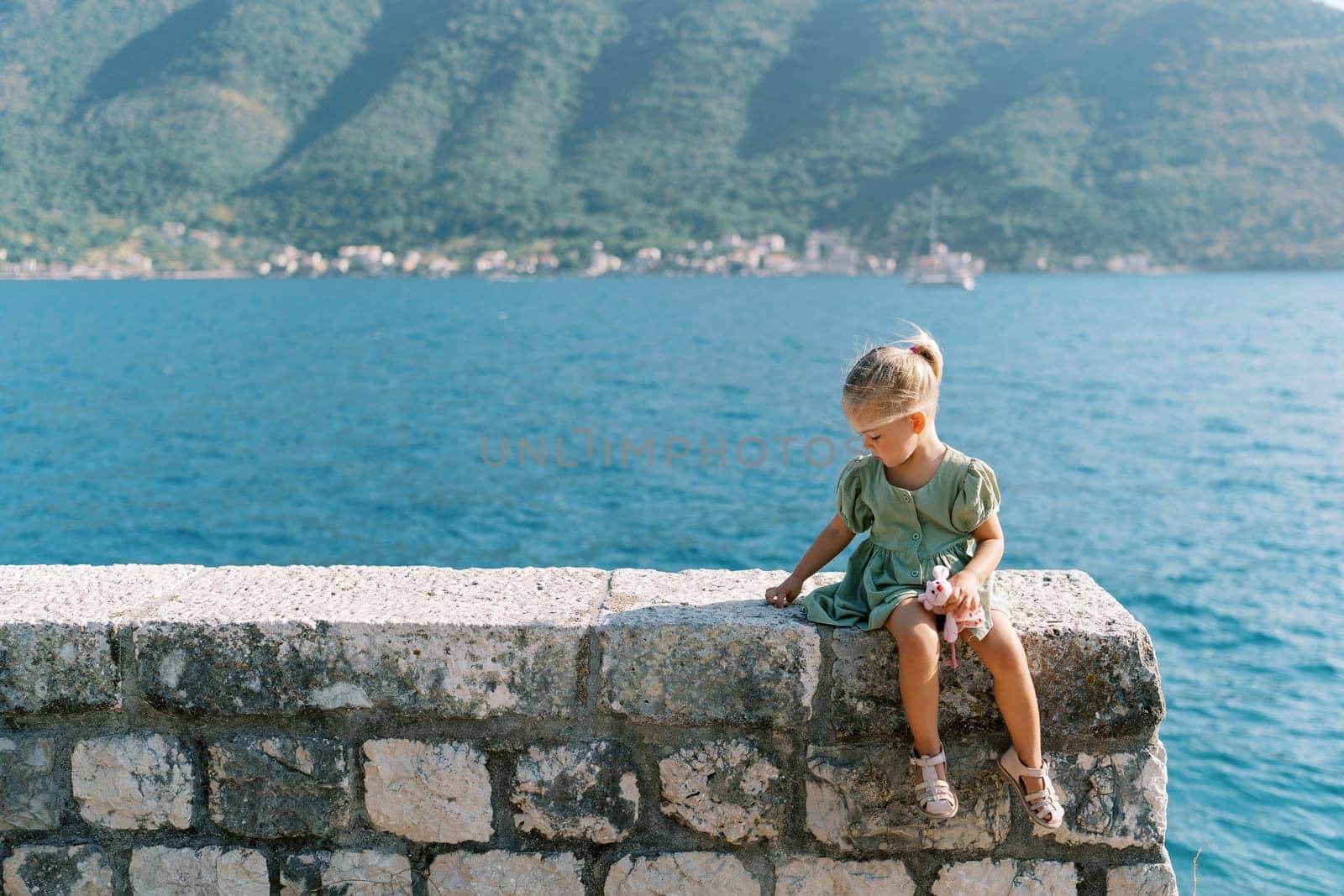 Little girl with a soft toy sits on a stone fence on the seashore and picks it with her finger. High quality photo