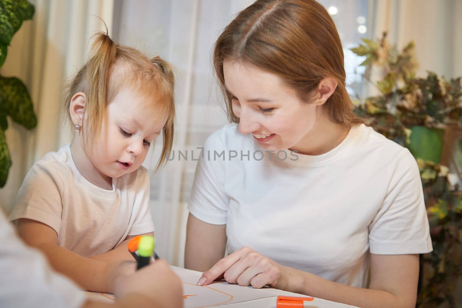 Young mother or babysitter, little daughter drawing at table in room. Painting, doing homework. Family enjoying leisure at home