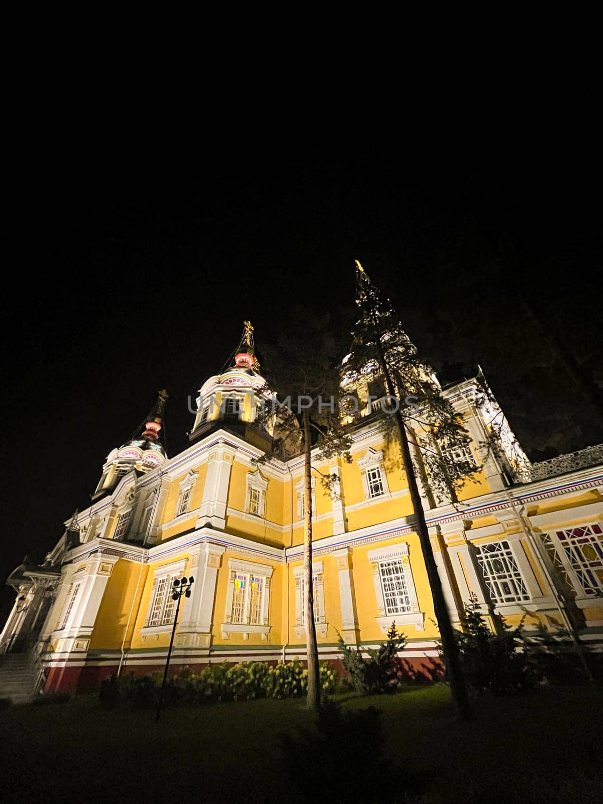 A stunning Russian Orthodox church illuminated at night, with a vibrant yellow exterior, towering steeple, surrounded by trees and park.