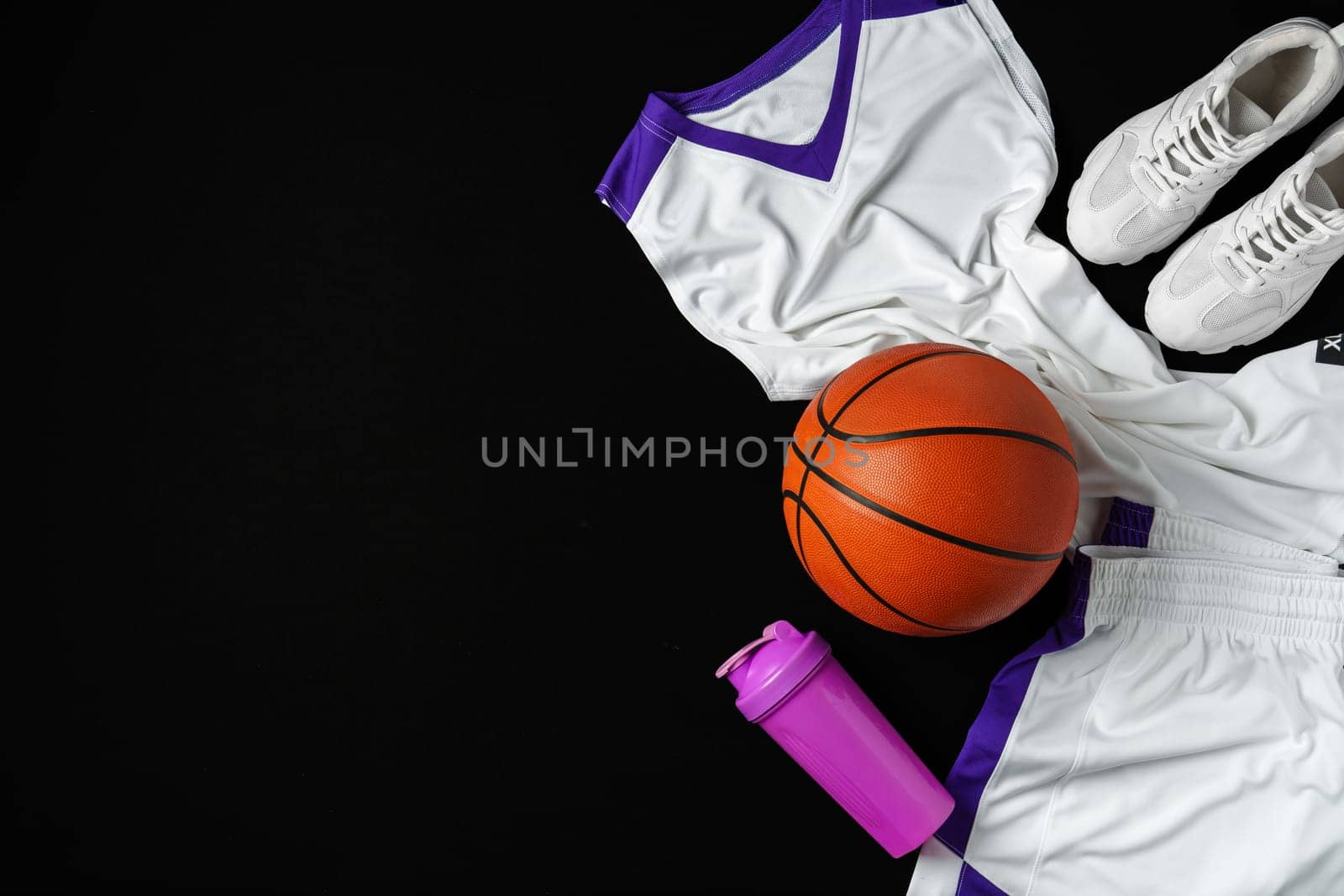 A collection of basketball essentials is neatly arranged against a stark, dark background, including a vibrant orange basketball, white sneakers with purple detailing, a white jersey with purple accents, and a purple water bottle, suggesting readiness for an athletic training session or a game.