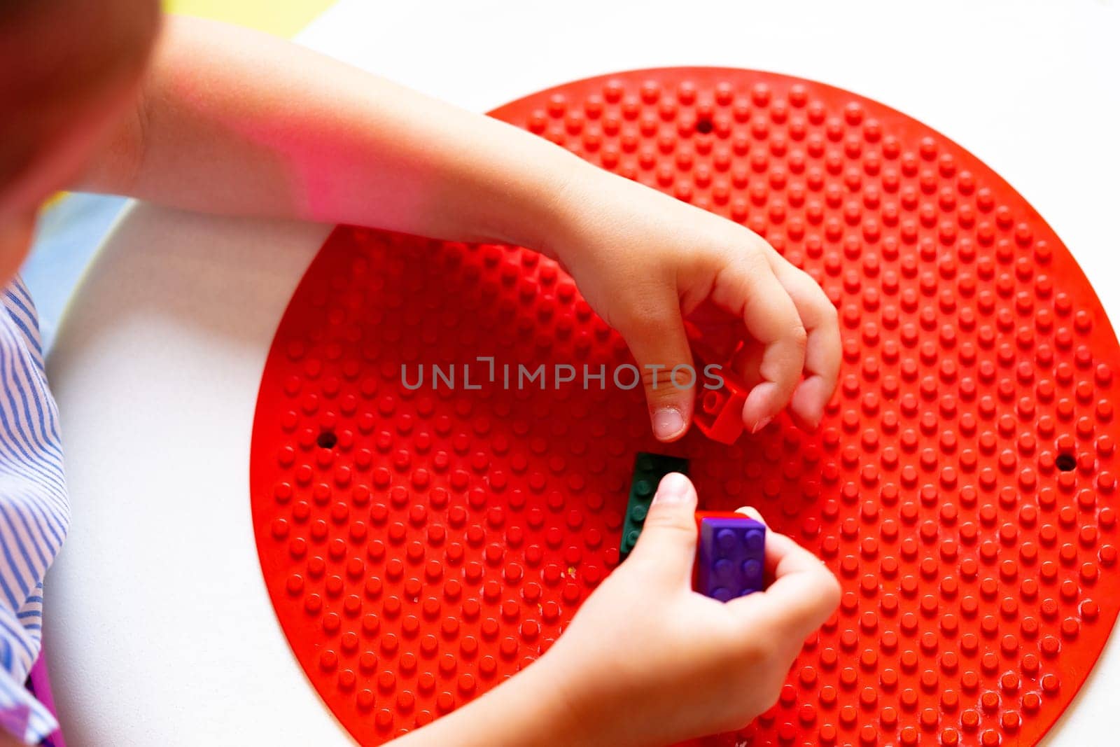 A young child is deeply focused while constructing a design with colorful plastic building blocks on a round red base. The childs hands are precisely placing a blue block to continue the pattern they have started, indicating an exercise in fine motor skills and creativity.
