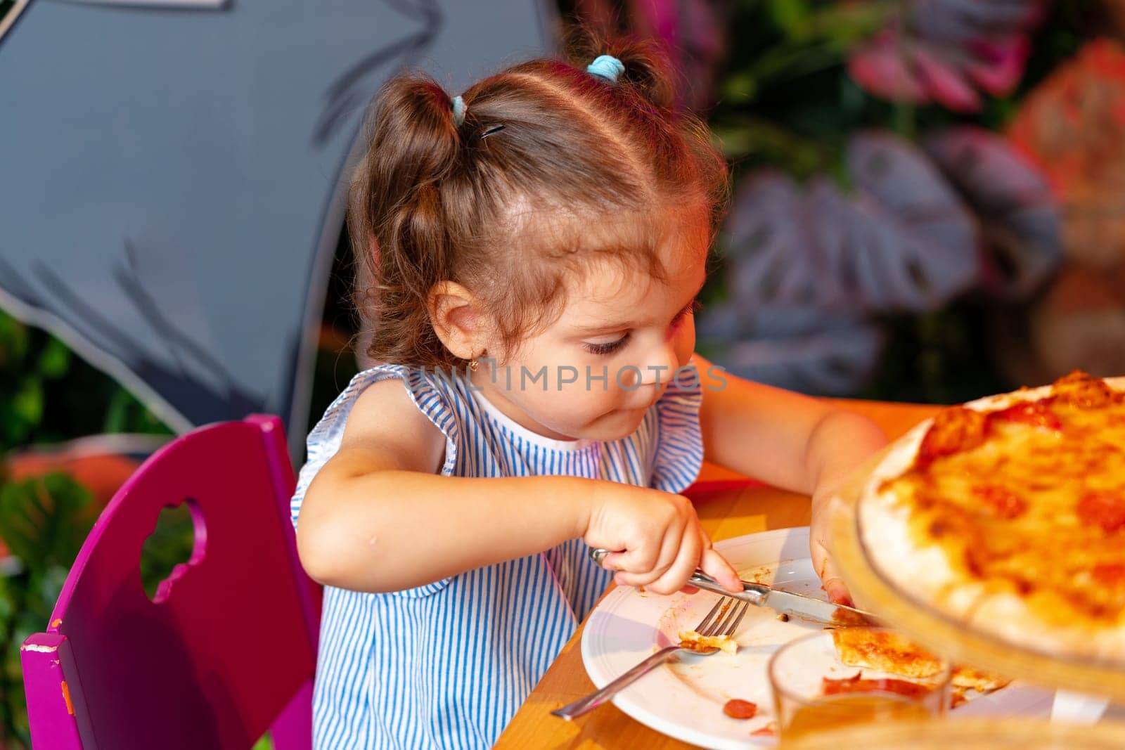 A young girl with light hair is captured in the moment of eating a slice of cheesy pizza, while another child is seated next to her, slightly out of focus. The scene suggests a casual birthday party setting with children enjoying food.