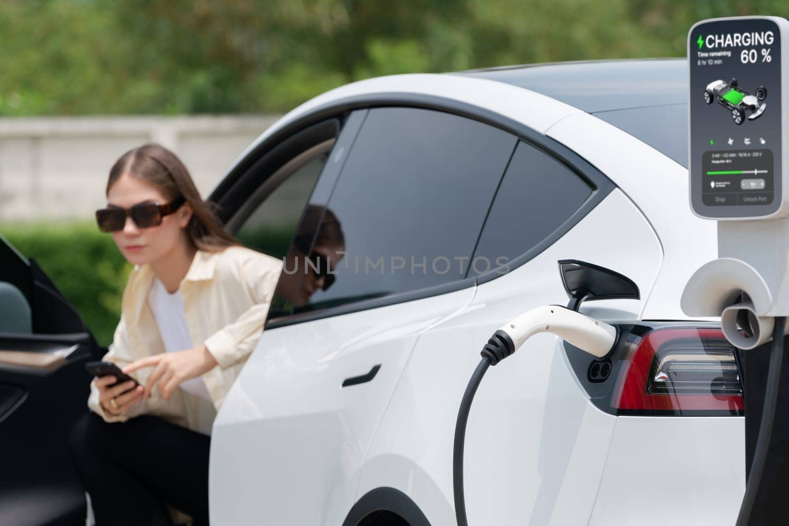 Young woman use smartphone to pay for electricity at public EV car charging station green city park. Modern environmental and sustainable urban lifestyle with EV vehicle. Expedient