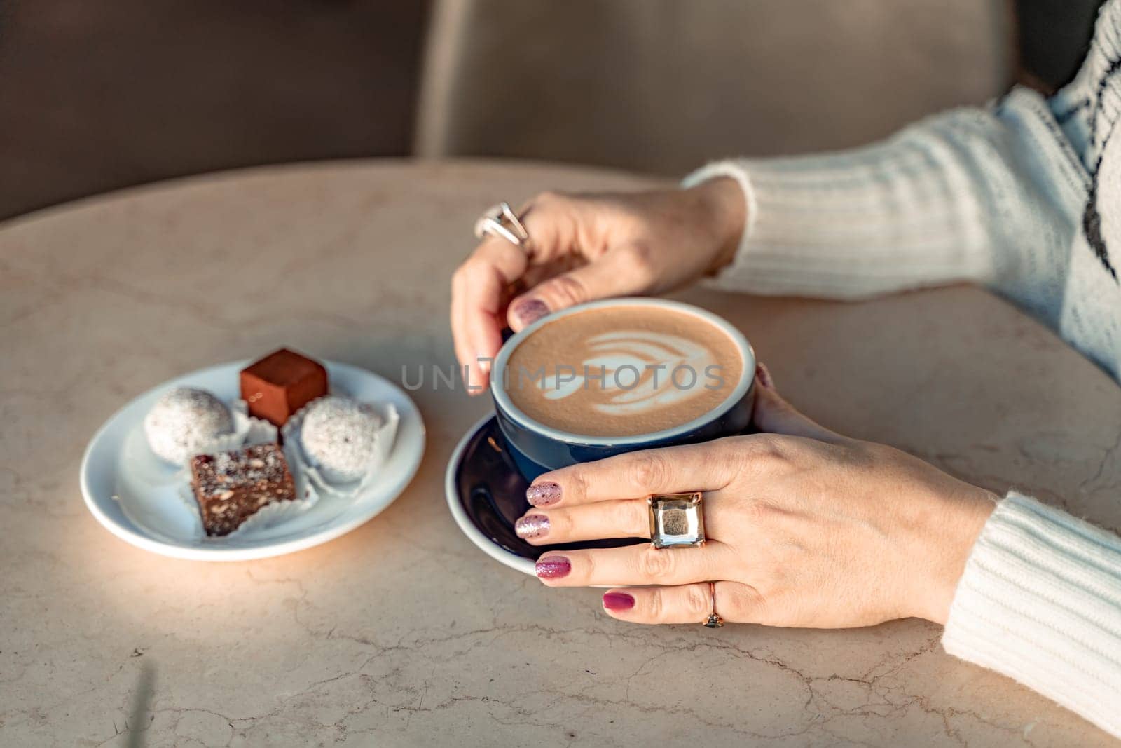 A cup of fresh cappuccino coffee in the hands of a woman on a fashionable background of a white marble table, next to a plate with sweets. Coffee addiction by Matiunina