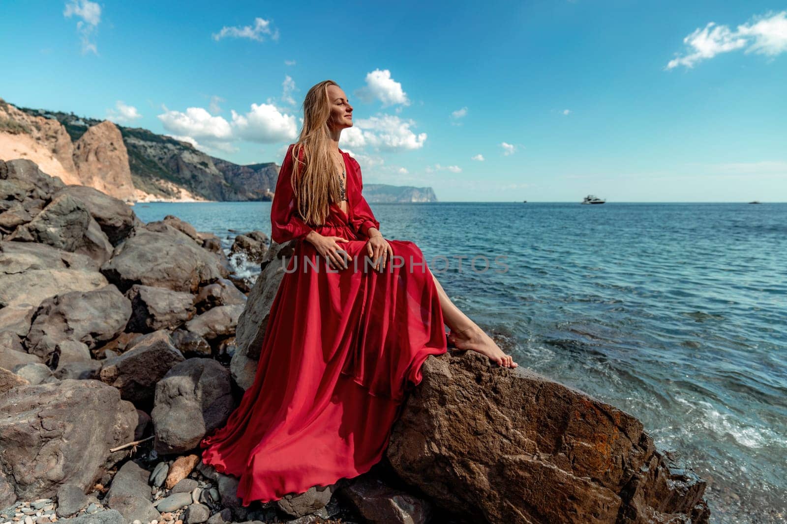 Red dress sea woman. A blonde with flowing hair in a long red dress sits on a rock near the sea. The concept of trips, a photo shoot at the sea.