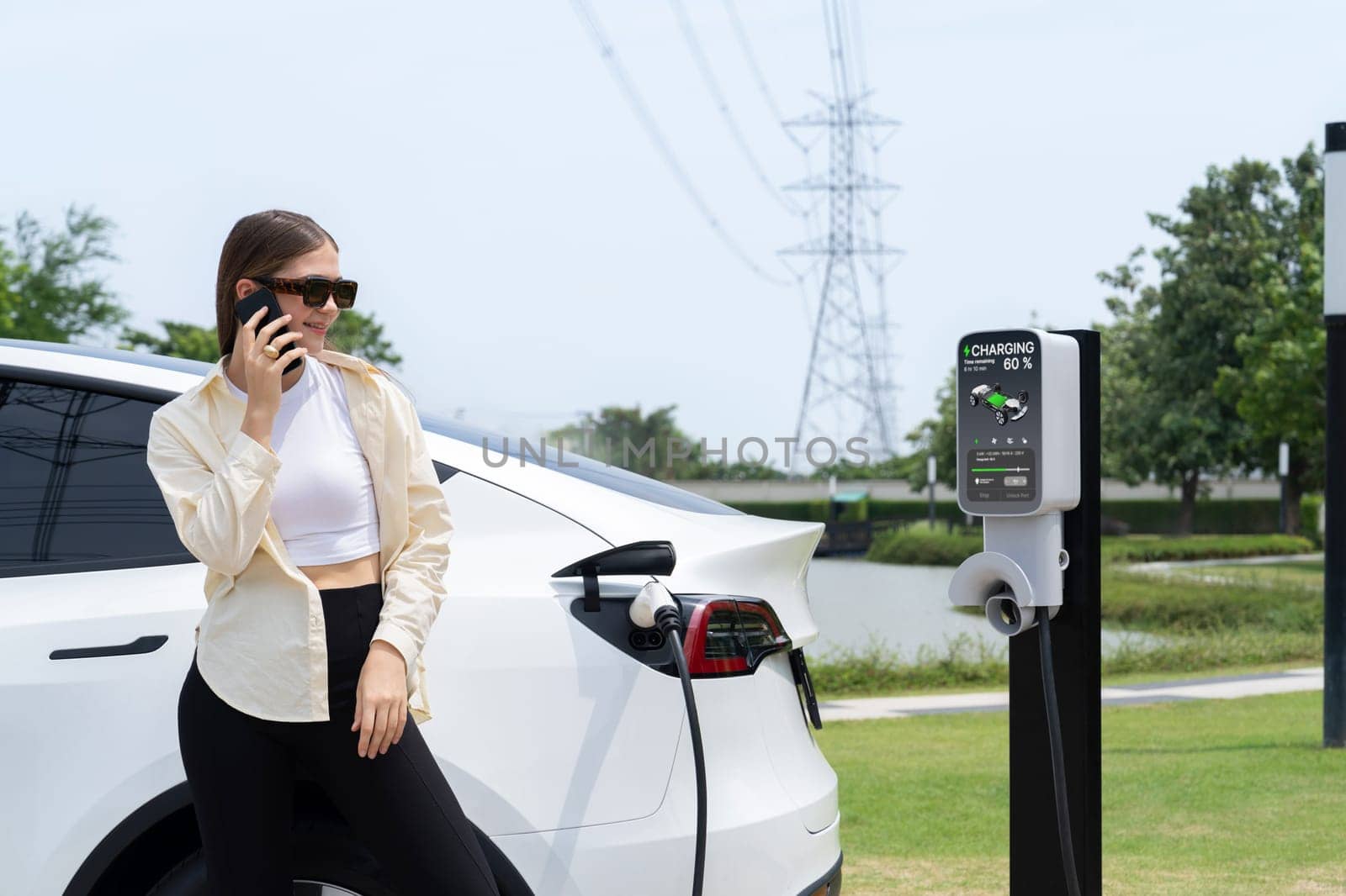 Young woman recharging EV car battery while talk on phone at charging station connected to electrical power grid tower facility as electrical industry for eco friendly vehicle utilization. Expedient