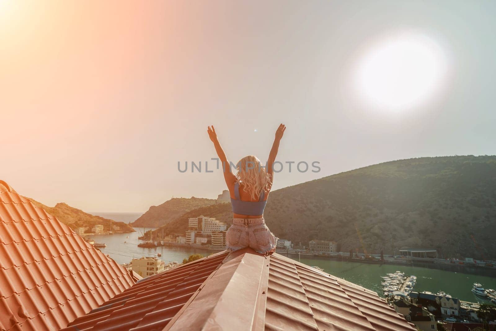 Woman sits on rooftop with outstretched arms, enjoys town view and sea mountains. Peaceful rooftop relaxation. Below her, there is a town with several boats visible in the water by Matiunina