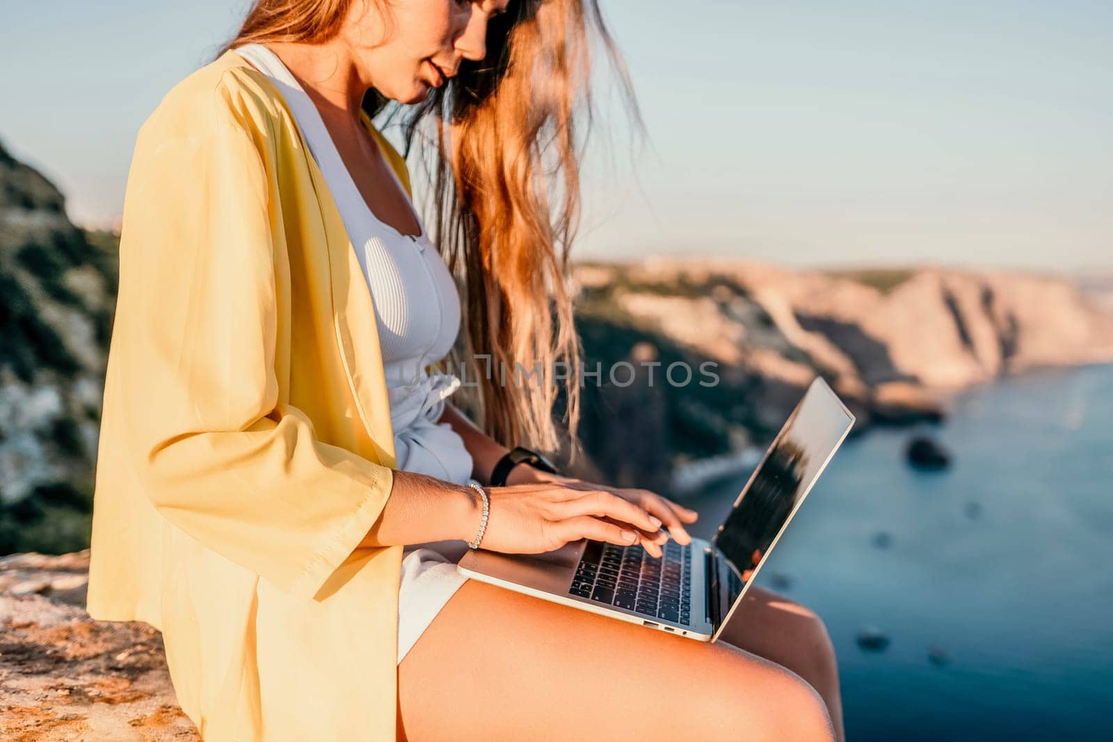 Successful business woman in yellow hat working on laptop by the sea. Pretty lady typing on computer at summer day outdoors. Freelance, travel and holidays concept.