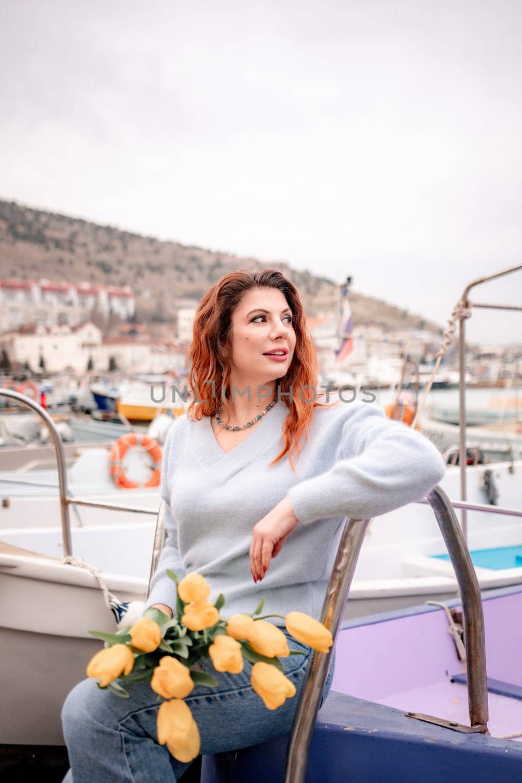 Woman holds yellow tulips in harbor with boats docked in the background., overcast day, yellow sweater, mountains.