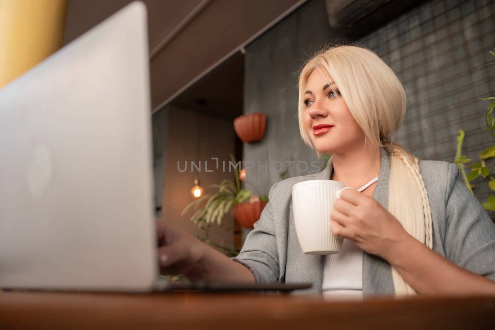 A blonde woman is sitting at a table with a laptop and a white coffee cup. She is wearing a gray jacket and has red lipstick on. The scene suggests a casual and relaxed atmosphere