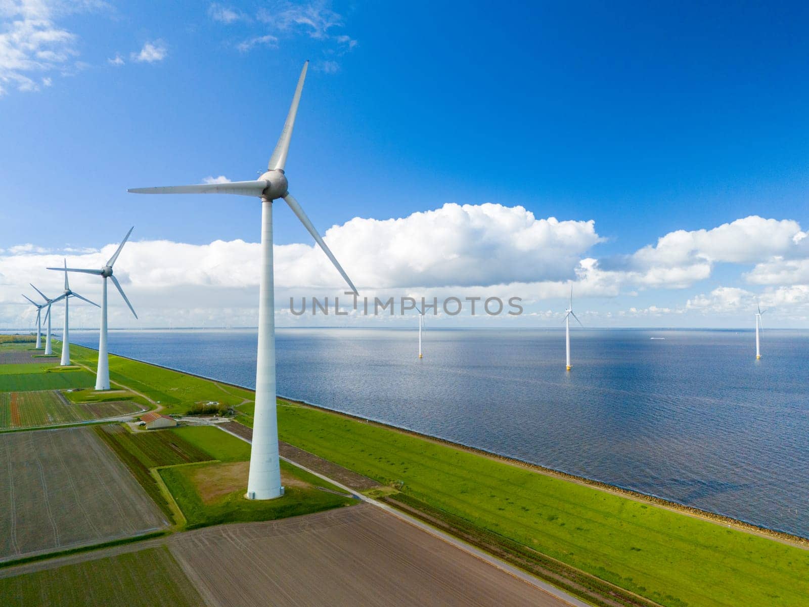 A wind farm composed of turbines stretches along the shore of a peaceful body of water in the Netherlands during the vibrant season of Spring by fokkebok