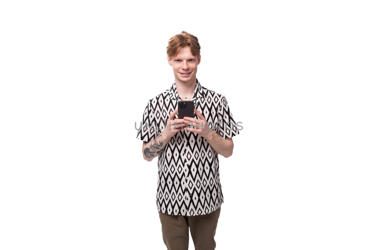 a young red-haired man in a summer shirt with a rhombus print is chatting in a smartphone against the background with copy space by TRMK