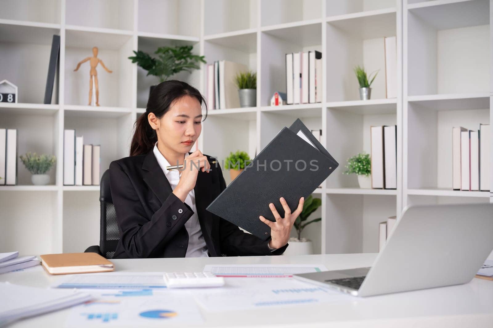 Focused Asian female accountant doing paperwork in office with plan documents on desk by wichayada