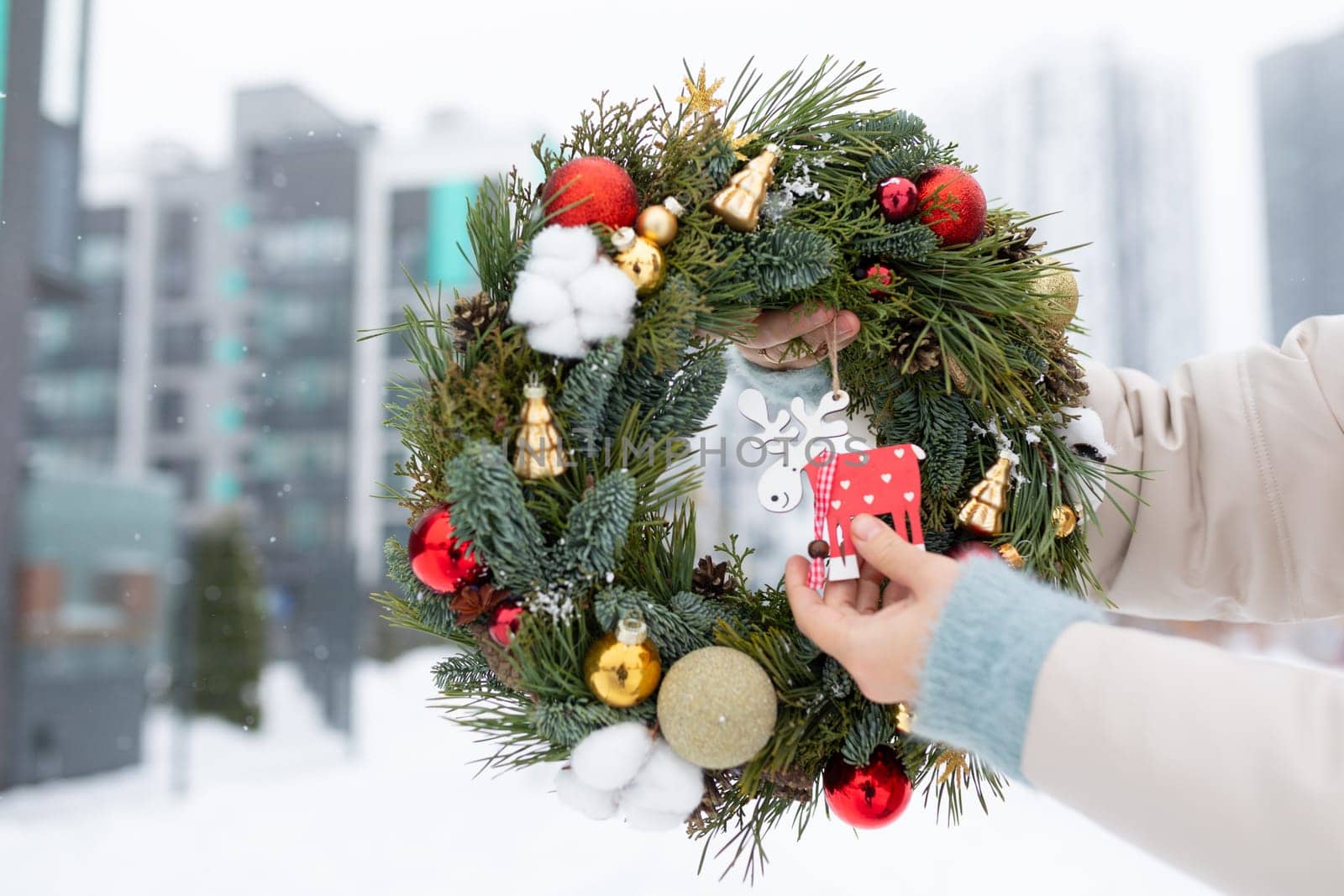 A person is holding a festive Christmas wreath adorned with colorful decorations. The wreath features ornaments, ribbons, and twinkling lights, creating a holiday atmosphere.