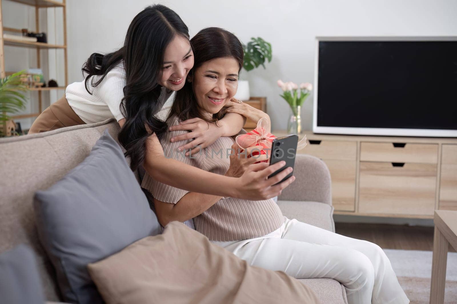 An elderly mother and her beautiful daughter happily lounge and take selfies together on the living room sofa..