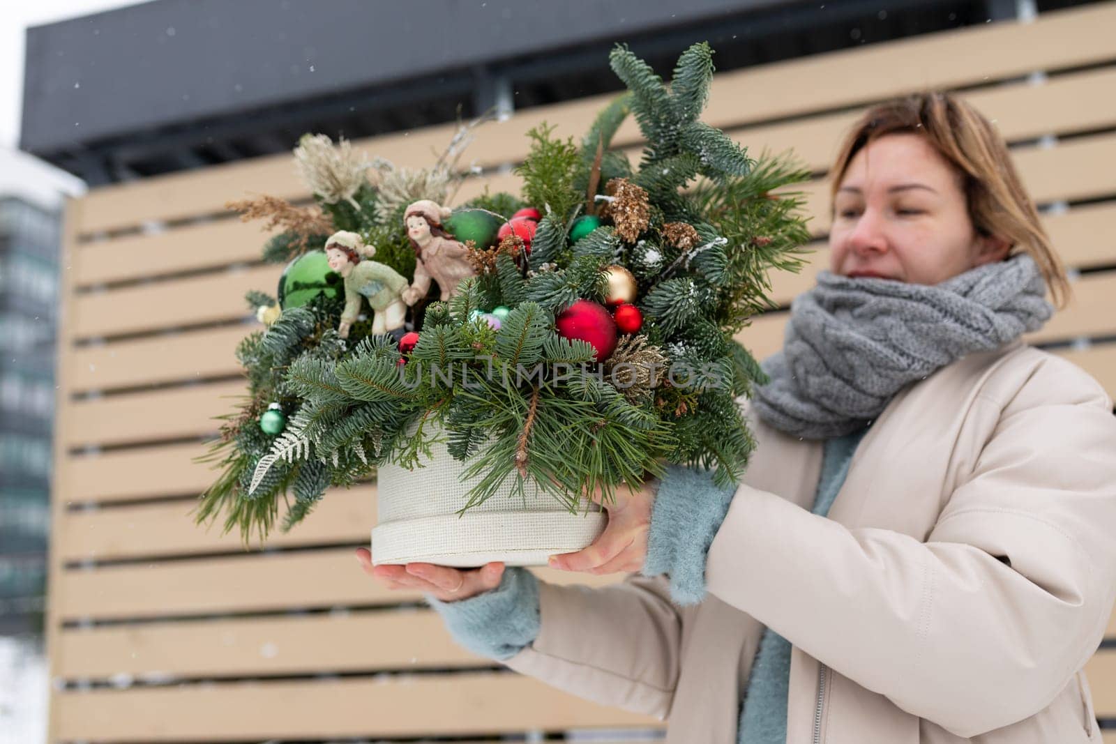 A woman is holding a potted plant decorated with festive Christmas ornaments. She is standing indoors, showcasing the seasonal display of greenery and holiday decorations.