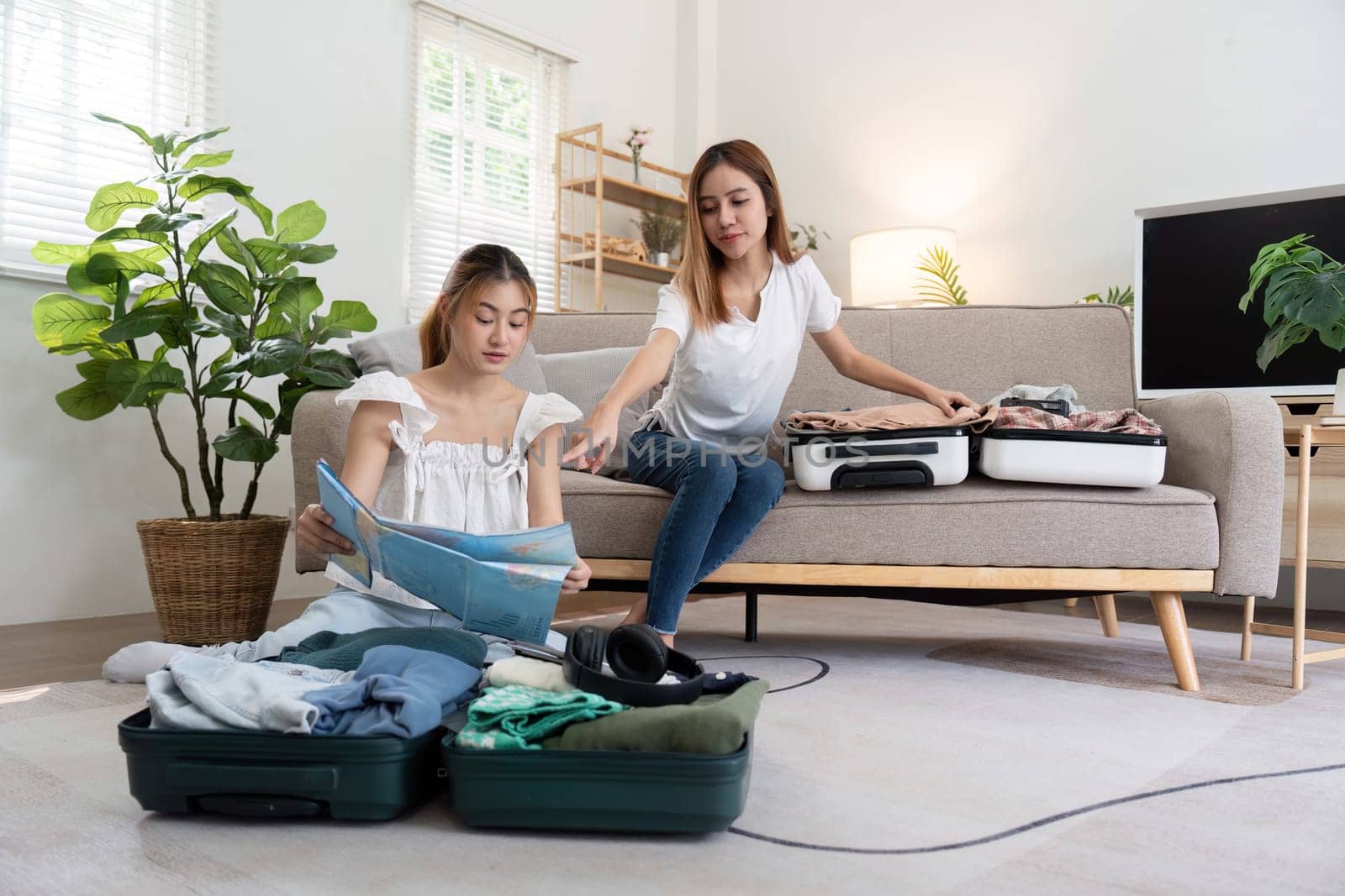 Young Asian woman packing clothes to the suitcase sitting on the sofa. Preparation for the summertime vacation. Two women are planning a trip and helping to prepare luggage to travel.