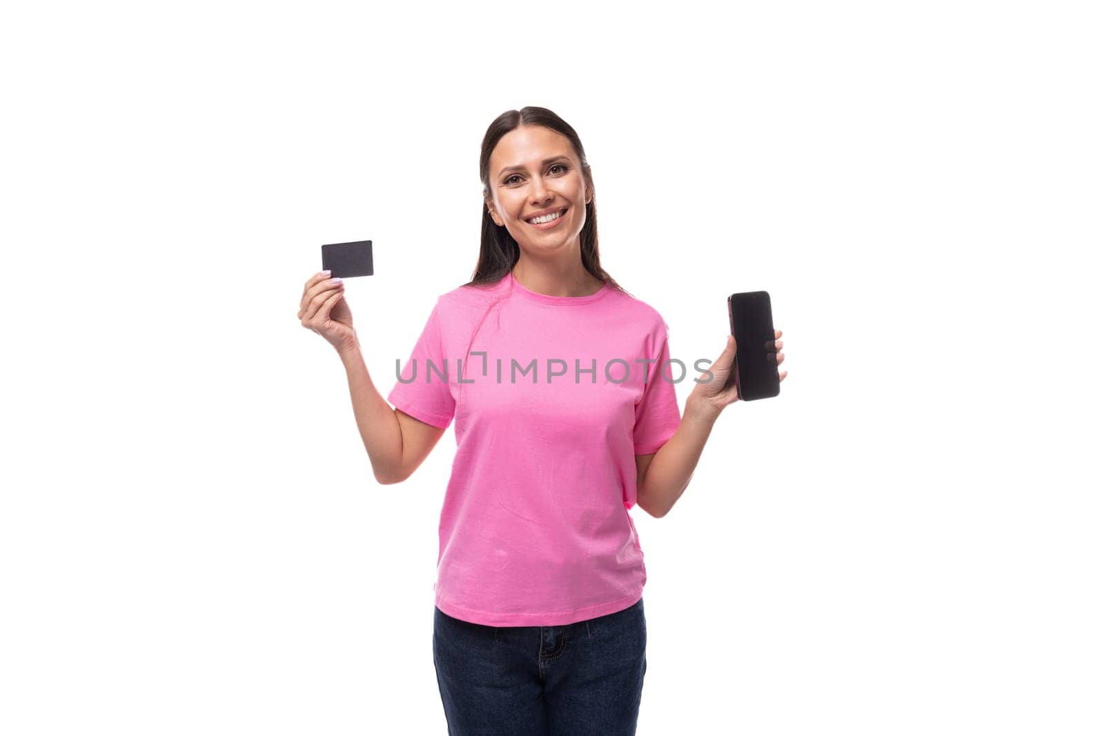 young smiling european woman with black hair in a pink t-shirt holding a smartphone with a mockup and a credit card on a white background with copy space.