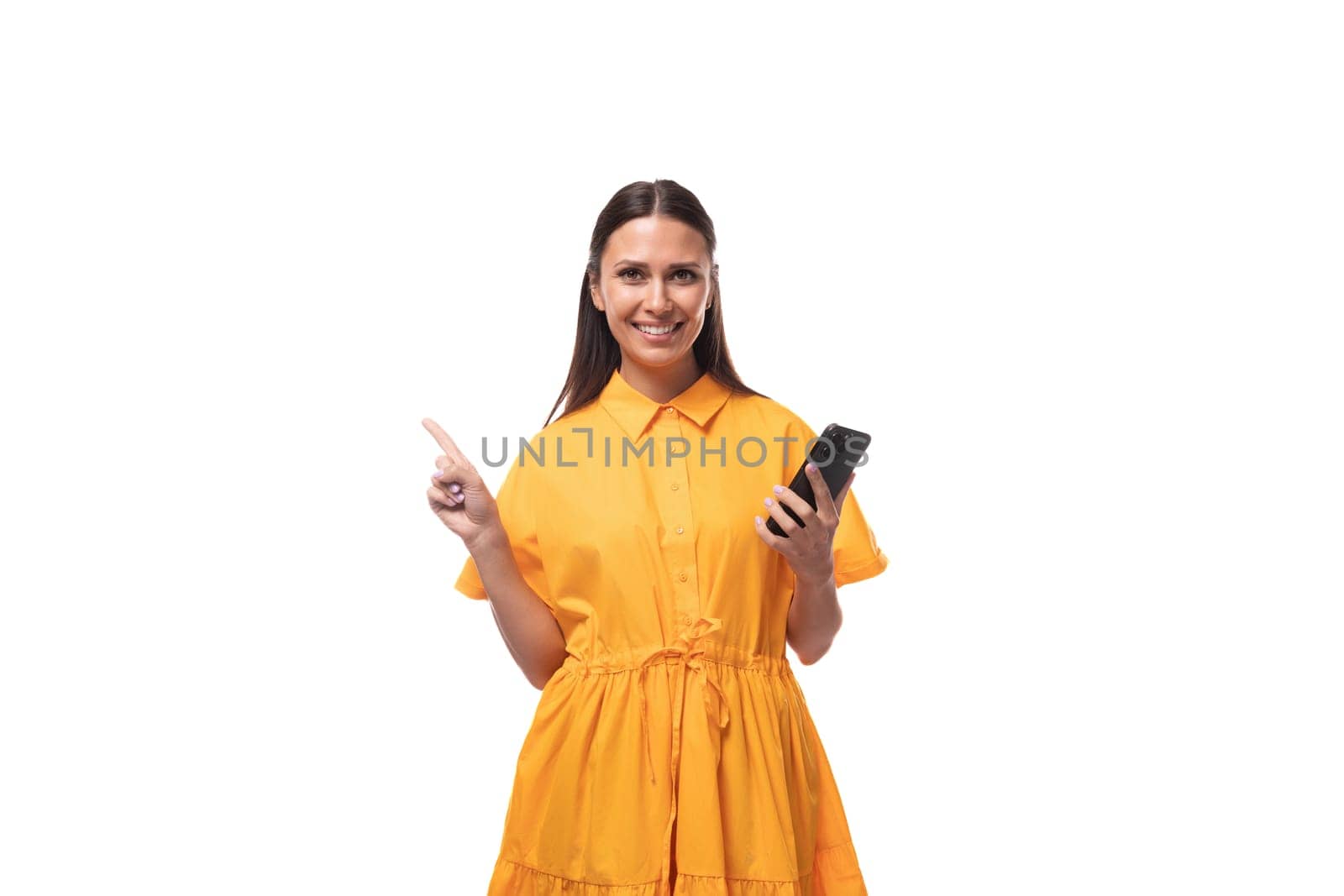 pretty young brunette lady dressed in a bright yellow dress on vacation smiling joyfully holding a phone in her hand.