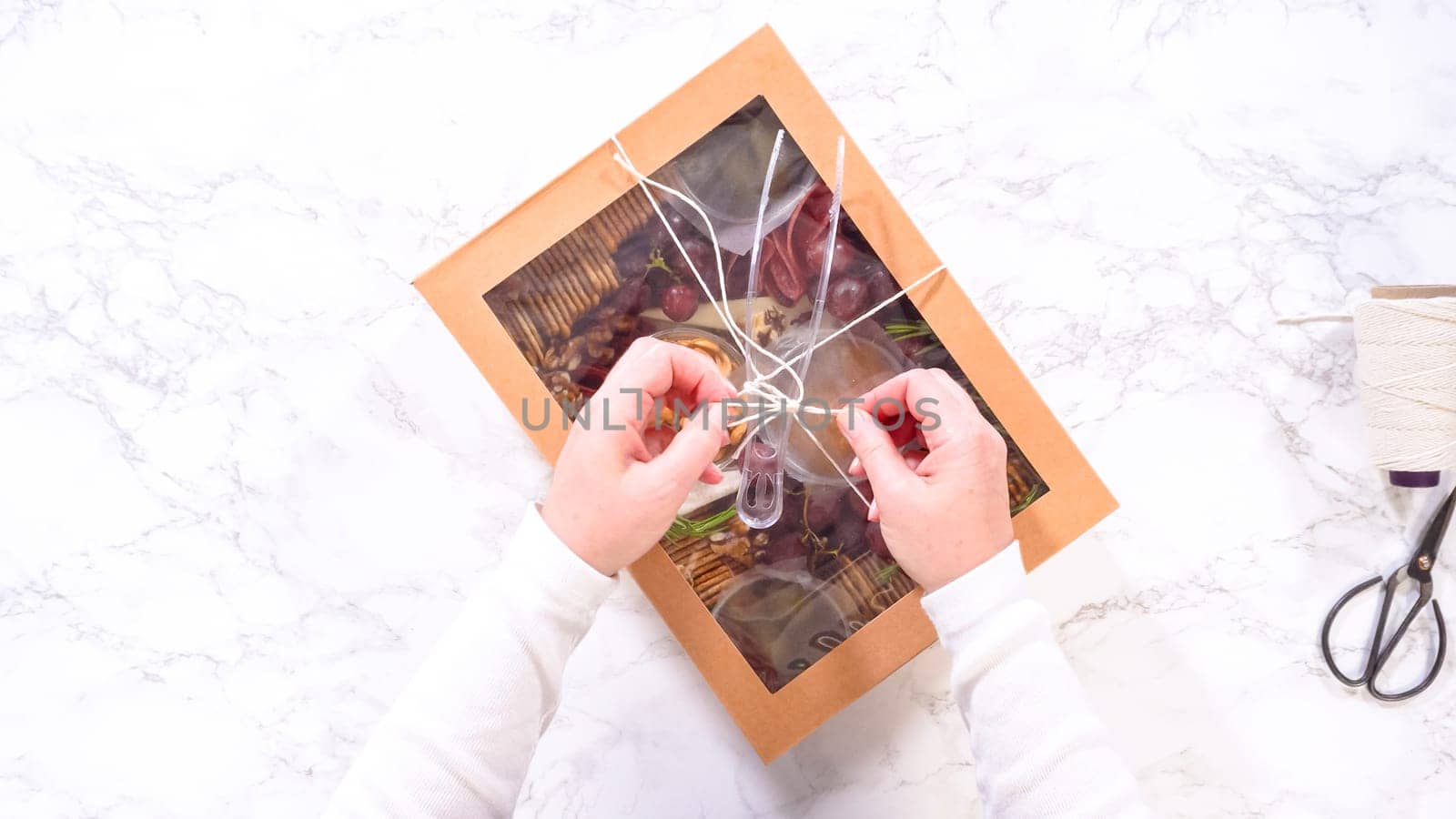 Flat lay. Hands are meticulously adding fresh red grapes to a bowl, complementing a beautifully arranged charcuterie box brimming with a variety of cheeses, olives, and cured meats, set against a sleek marble surface.